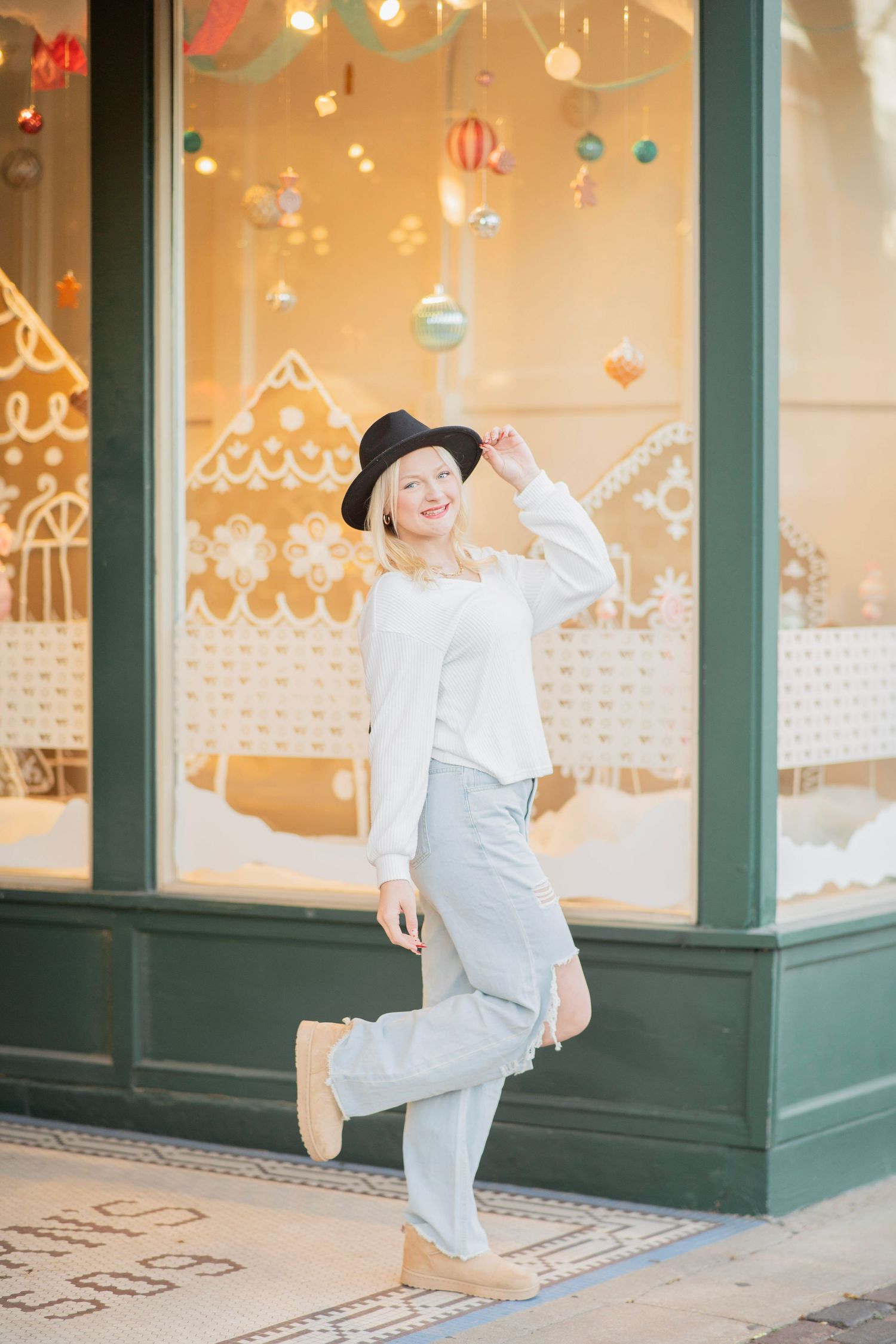 Stylish blonde woman in white top, gray pants and black hat poses outside festive holiday storefront.