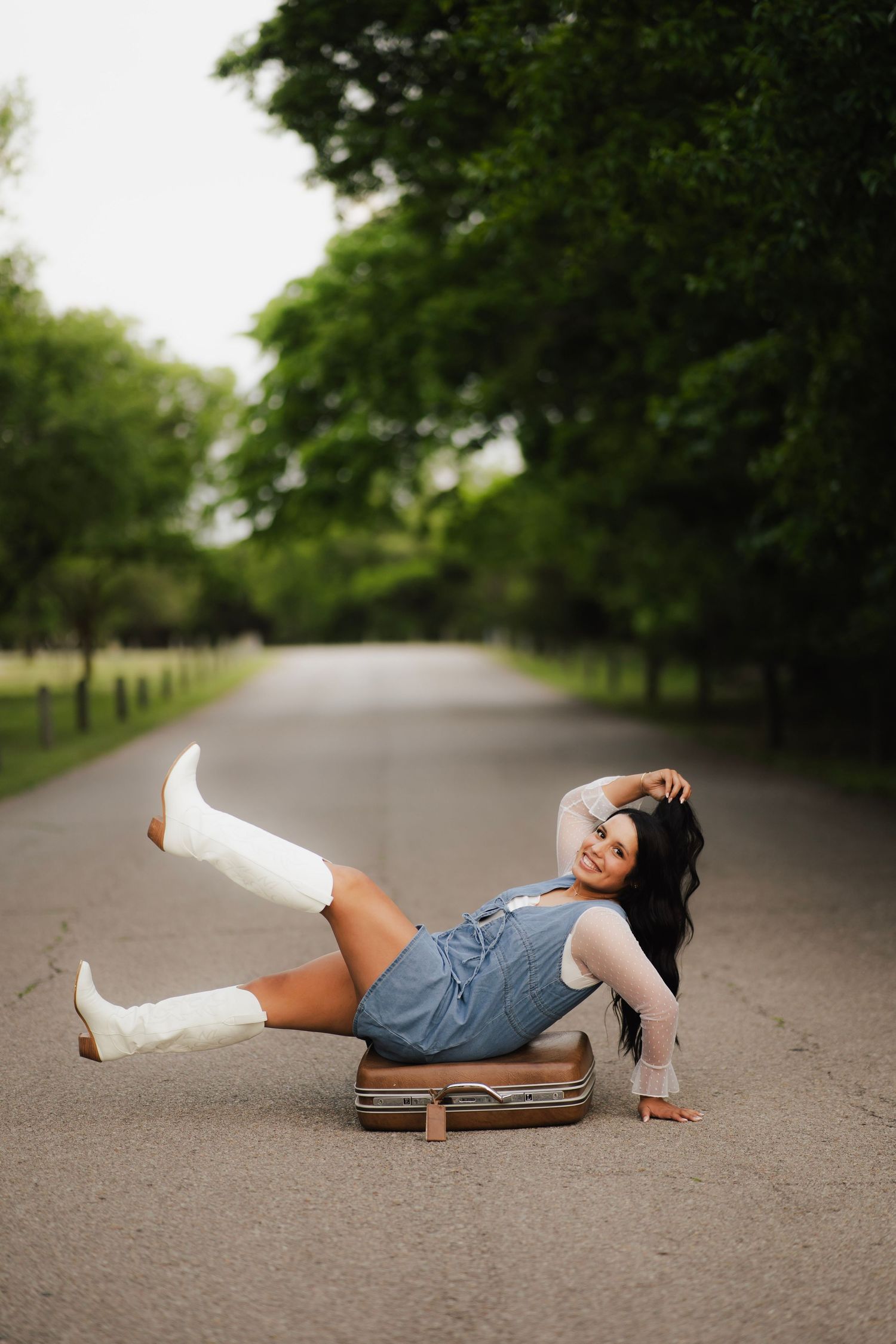 Young woman in denim dress and white cowboy boots sitting on vintage suitcase on a road.