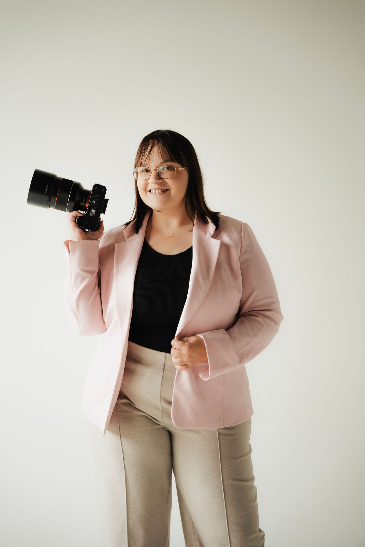 Smiling female photographer in pink blazer posing with professional DSLR camera on white background.