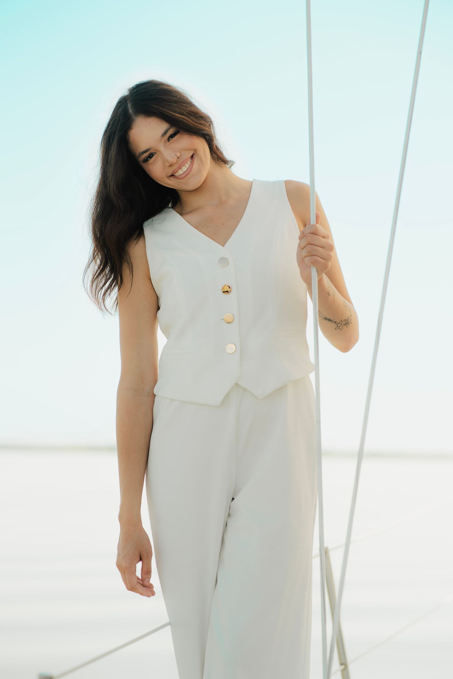 Smiling woman in white vest and trousers standing on a sailboat deck against a clear blue sky.