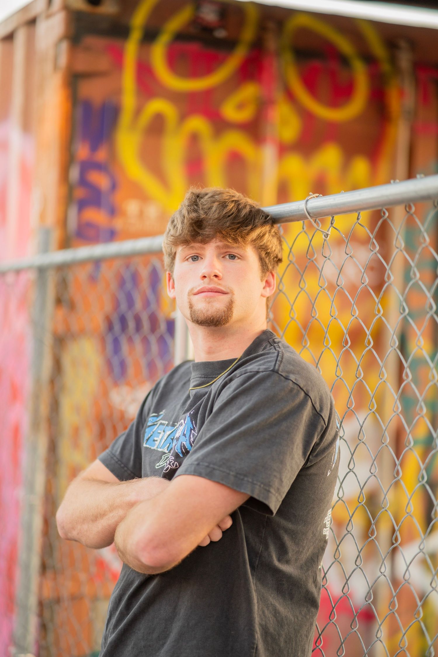 Young man with crossed arms leaning against chain-link fence with colorful graffiti wall backdrop.
