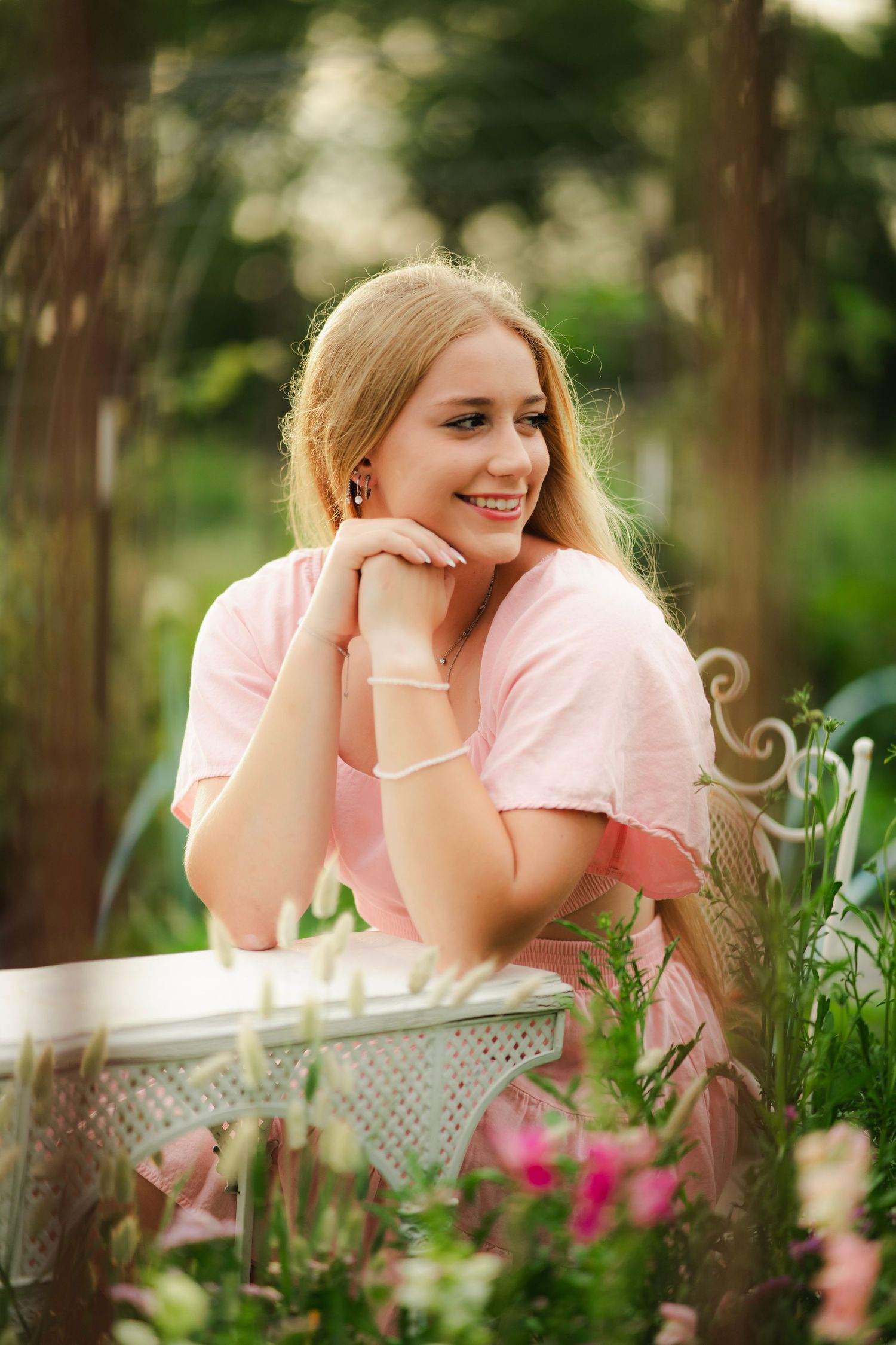 Smiling blonde woman in pink dress leaning on white table in garden setting.