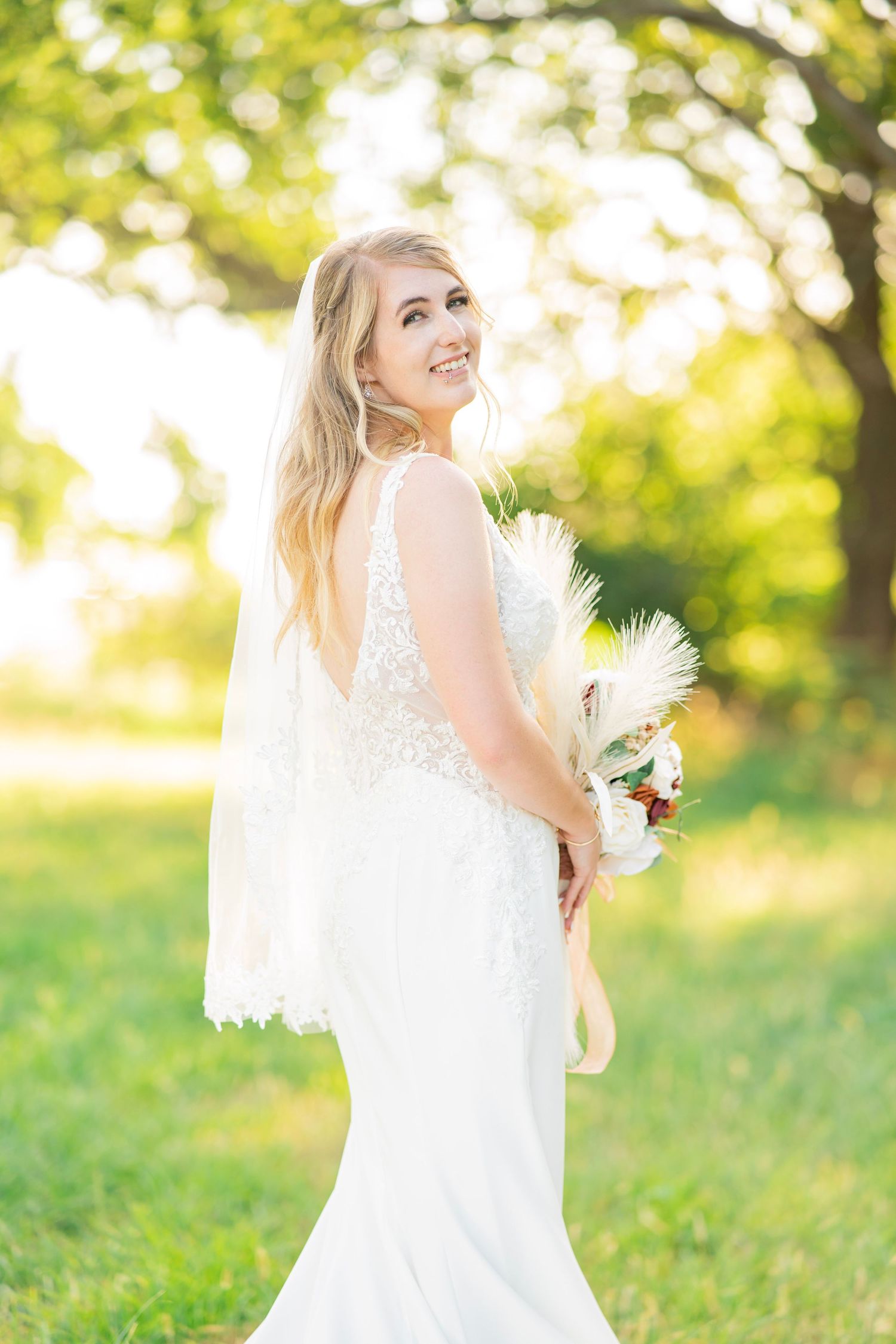 Smiling bride in lace wedding gown holding boho bouquet outdoors in golden sunlight green setting.
