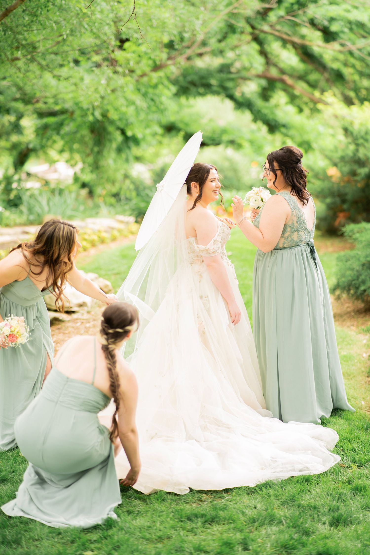 Bride in white gown and veil surrounded by bridesmaids in sage green dresses in a lush garden setting.