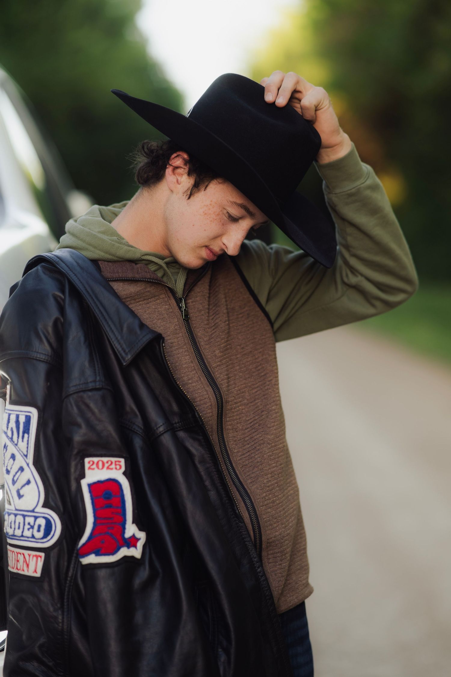 Young man in black cowboy hat and leather jacket with rodeo patches poses outdoors on a rural road.