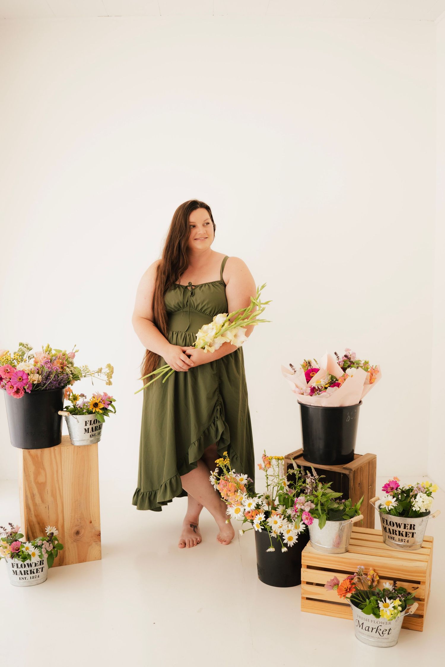 Woman in green dress holding flowers surrounded by flower market buckets and crates on white background.