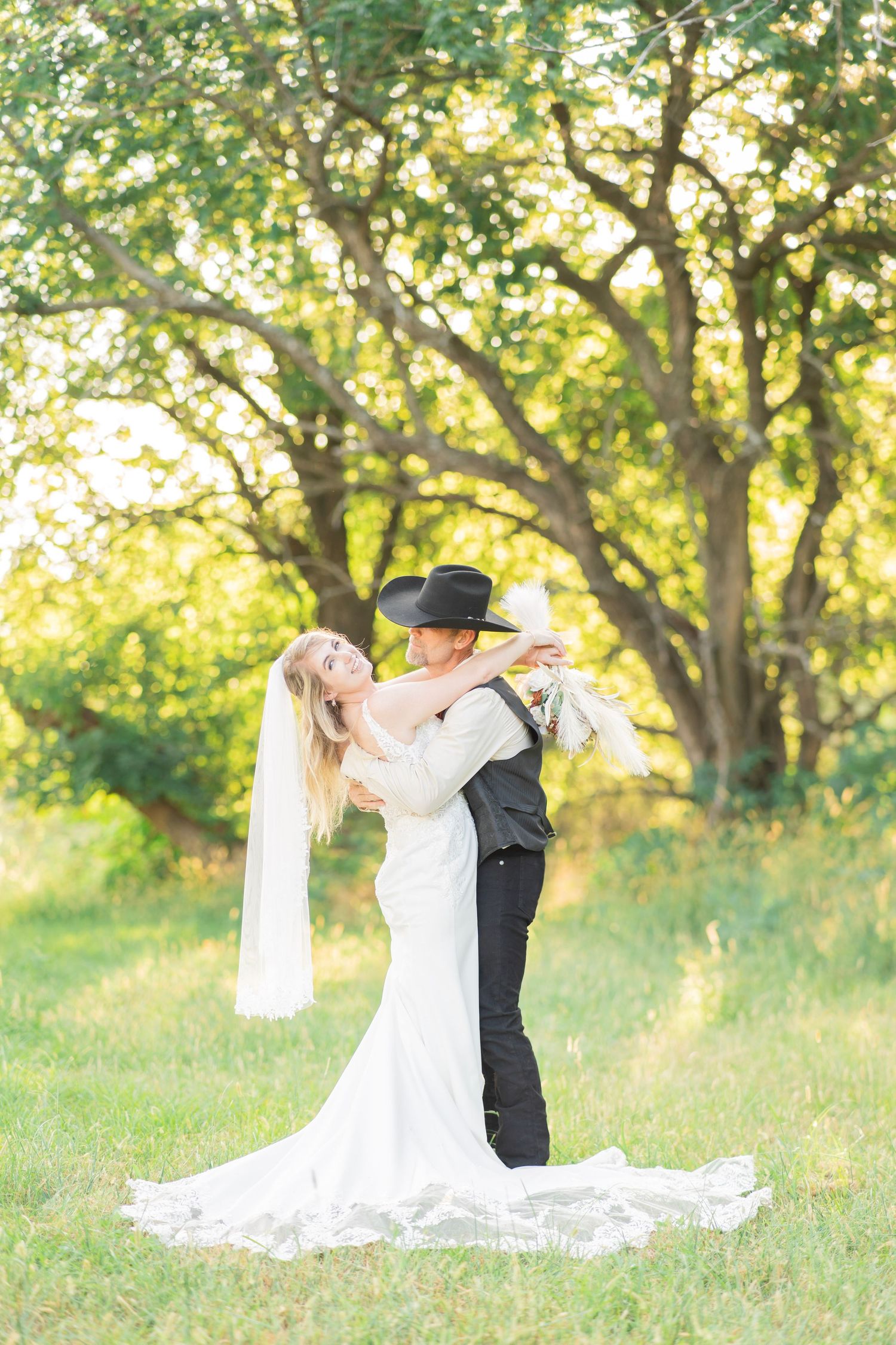 Bride and groom embracing outdoors, groom wearing black cowboy hat, bride in lace wedding dress.