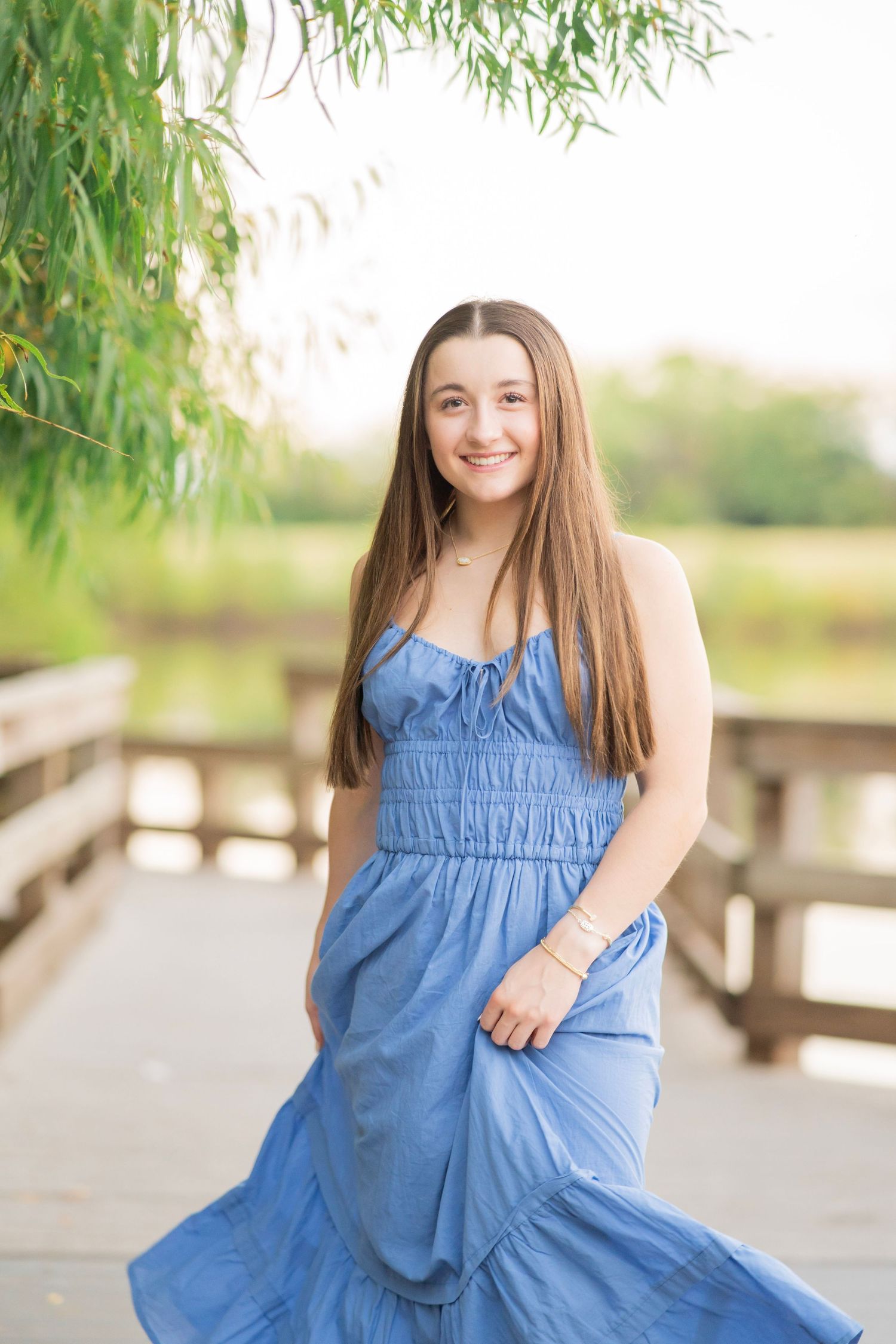Smiling teenage girl in blue maxi dress on wooden bridge surrounded by greenery.