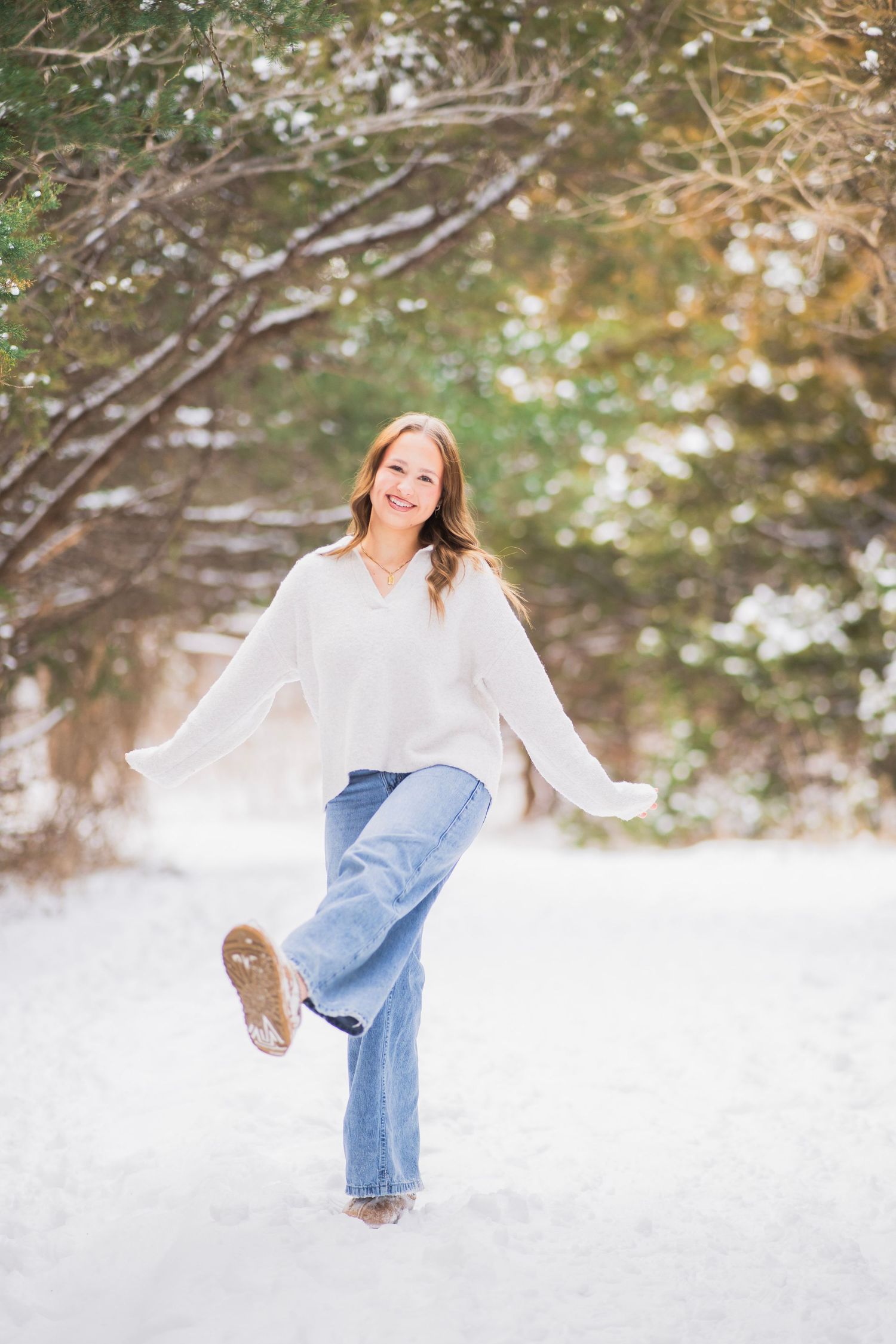 Young woman in white sweater and jeans playfully kicks her leg up in a snowy winter landscape.