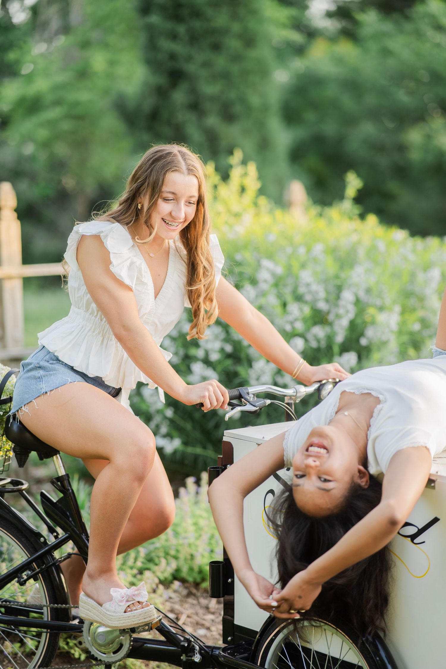 Two smiling young women enjoy a sunny day outdoors with a cargo bike surrounded by lush green gardens.
