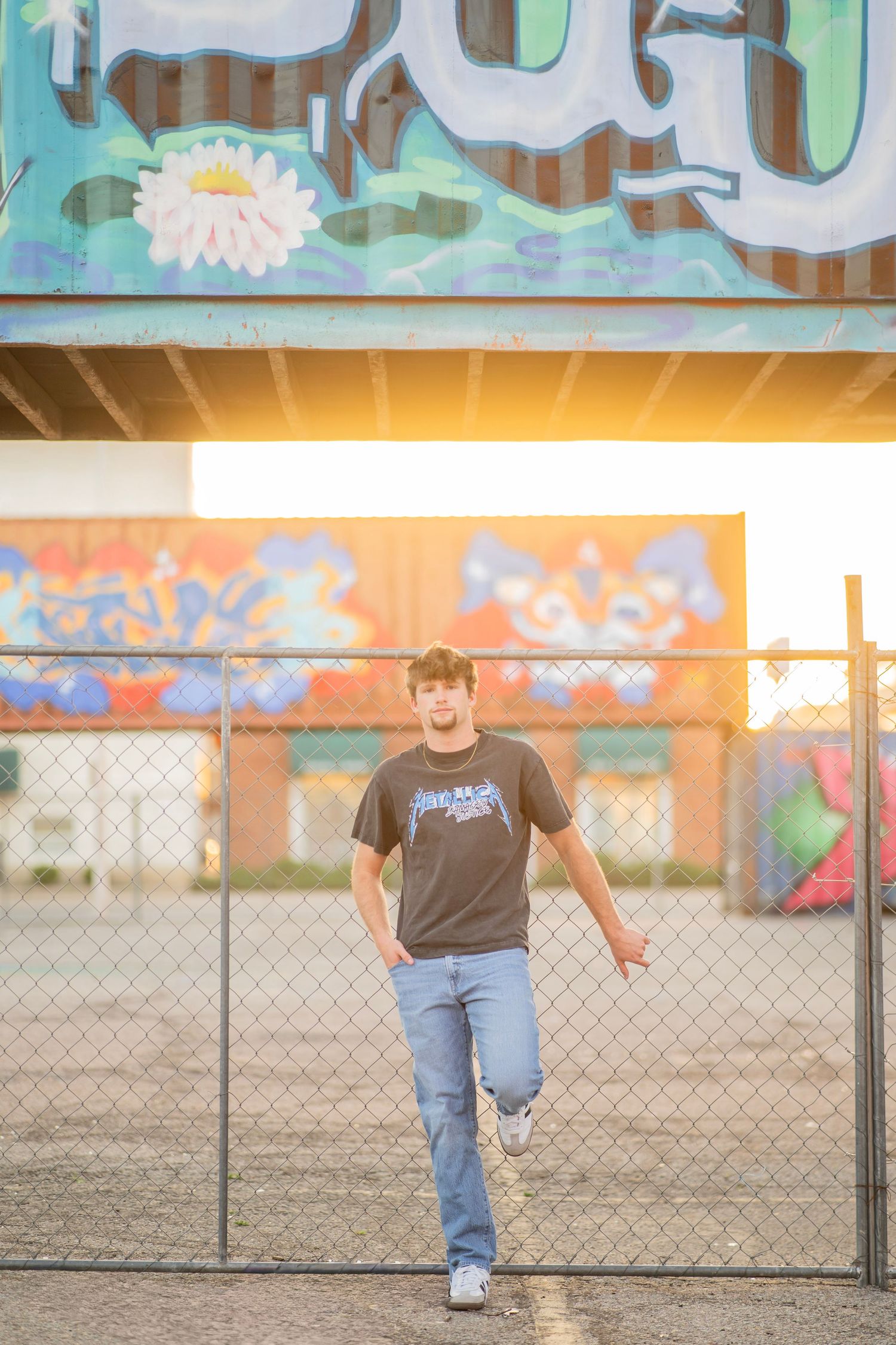 Young man in jeans and graphic tee leaning on chain-link fence with colorful graffiti murals and golden sunset backdrop.