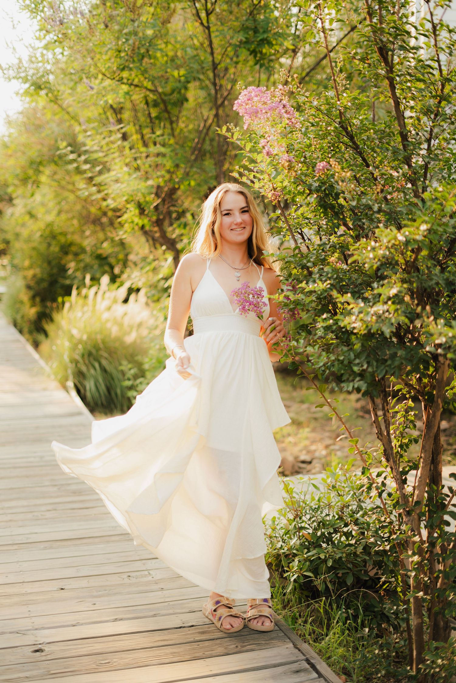 Smiling blonde woman in flowing white dress standing on wooden path surrounded by lush green trees and pink flowers.