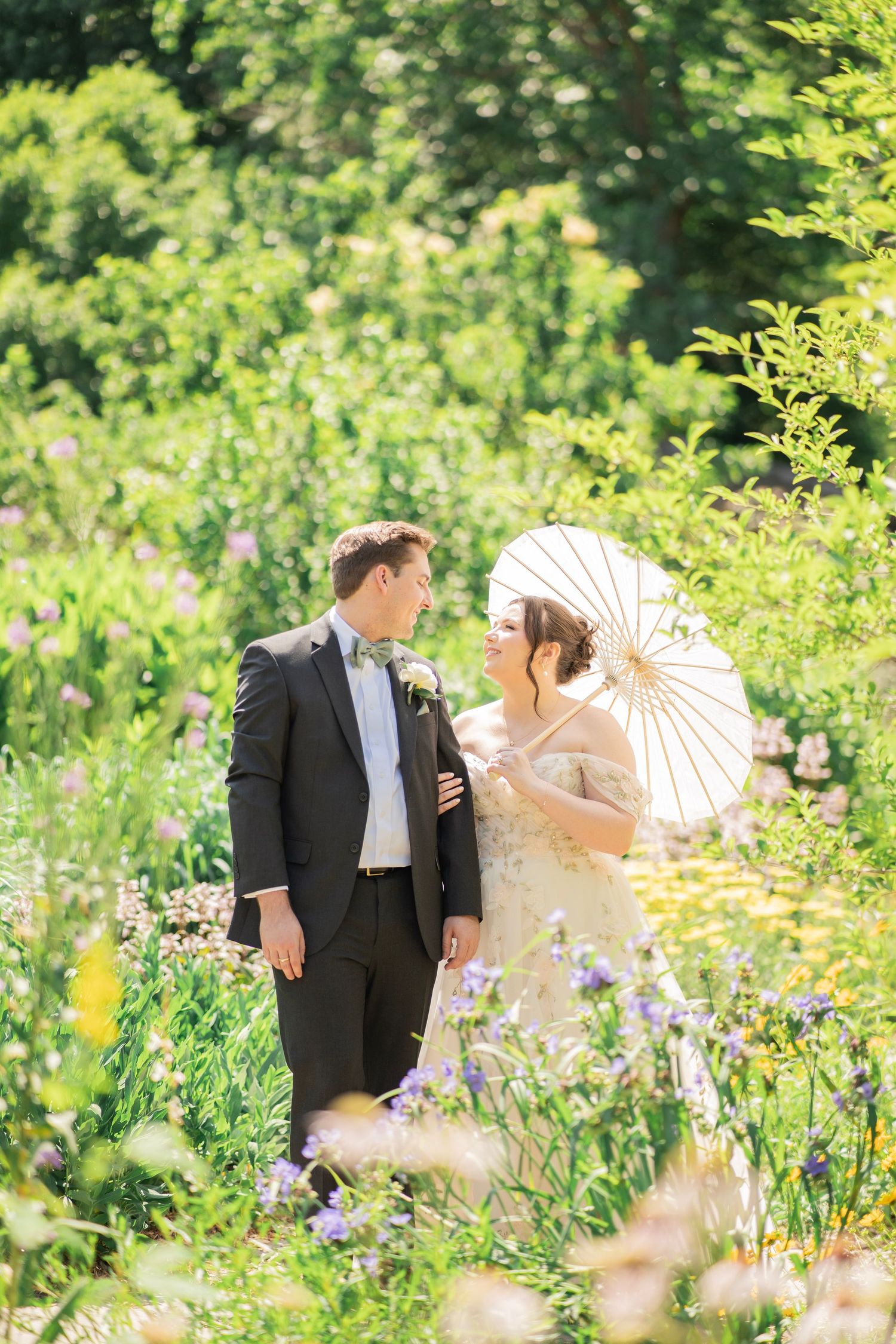 Bride holding white parasol and groom share a romantic moment surrounded by wildflowers in a lush garden.