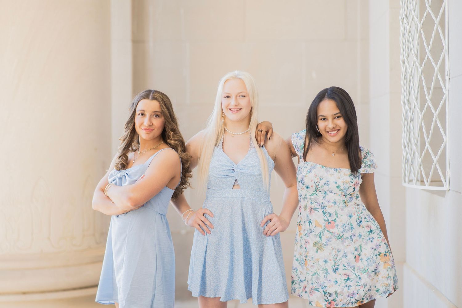 Three smiling young women in light pastel dresses posing together near elegant white columns.