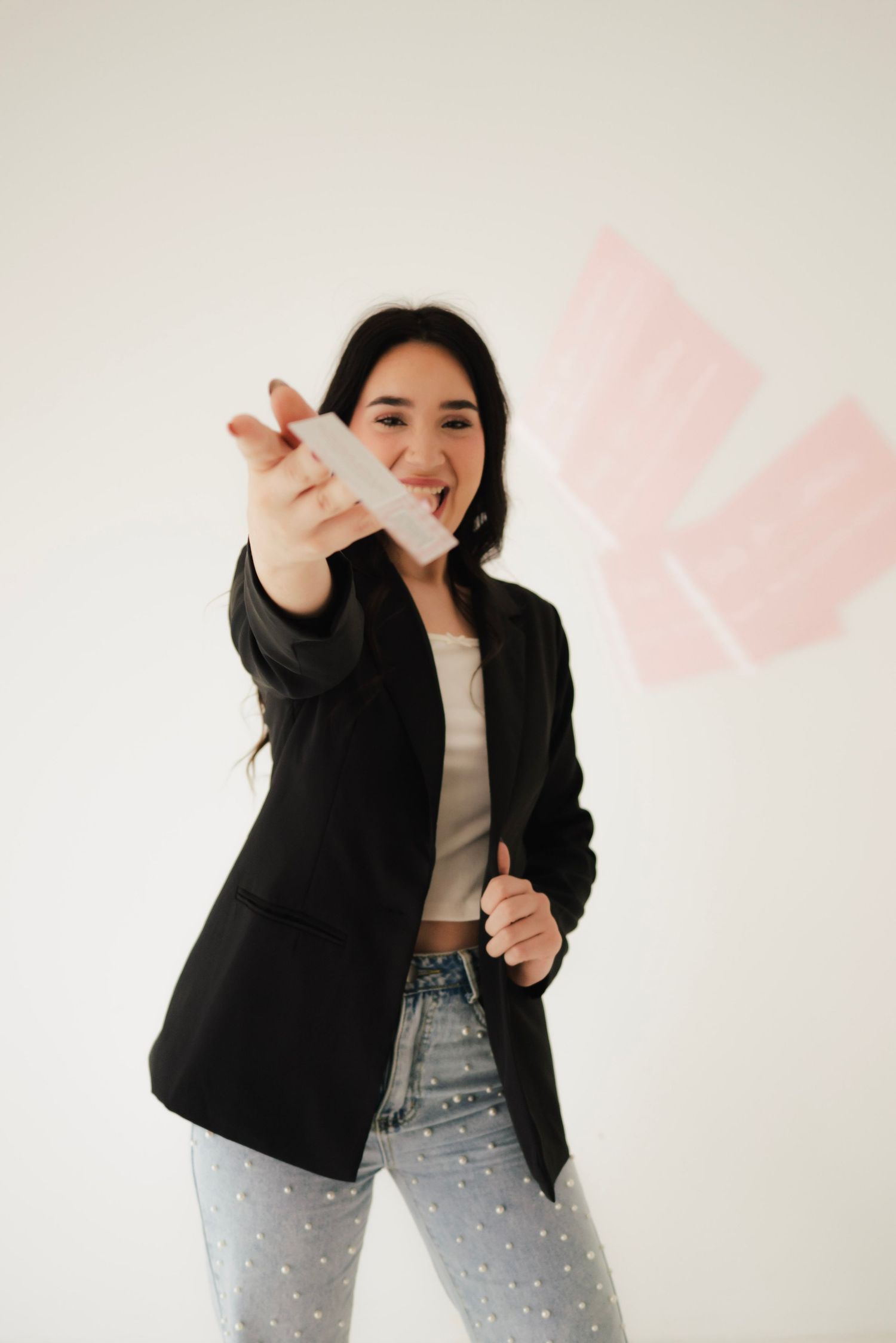 Young woman in black blazer and jeans smiling while holding object toward camera against white wall.