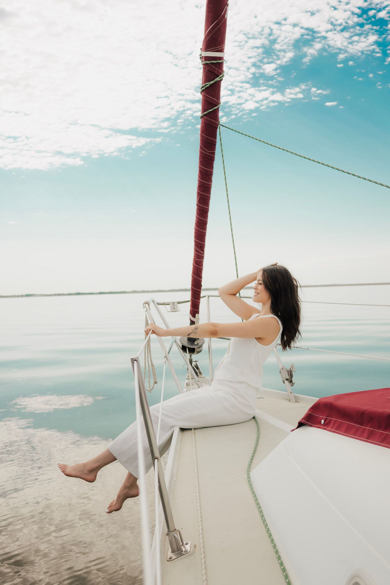 Woman in white outfit relaxing on sailboat deck, enjoying serene ocean views under a blue cloudy sky.