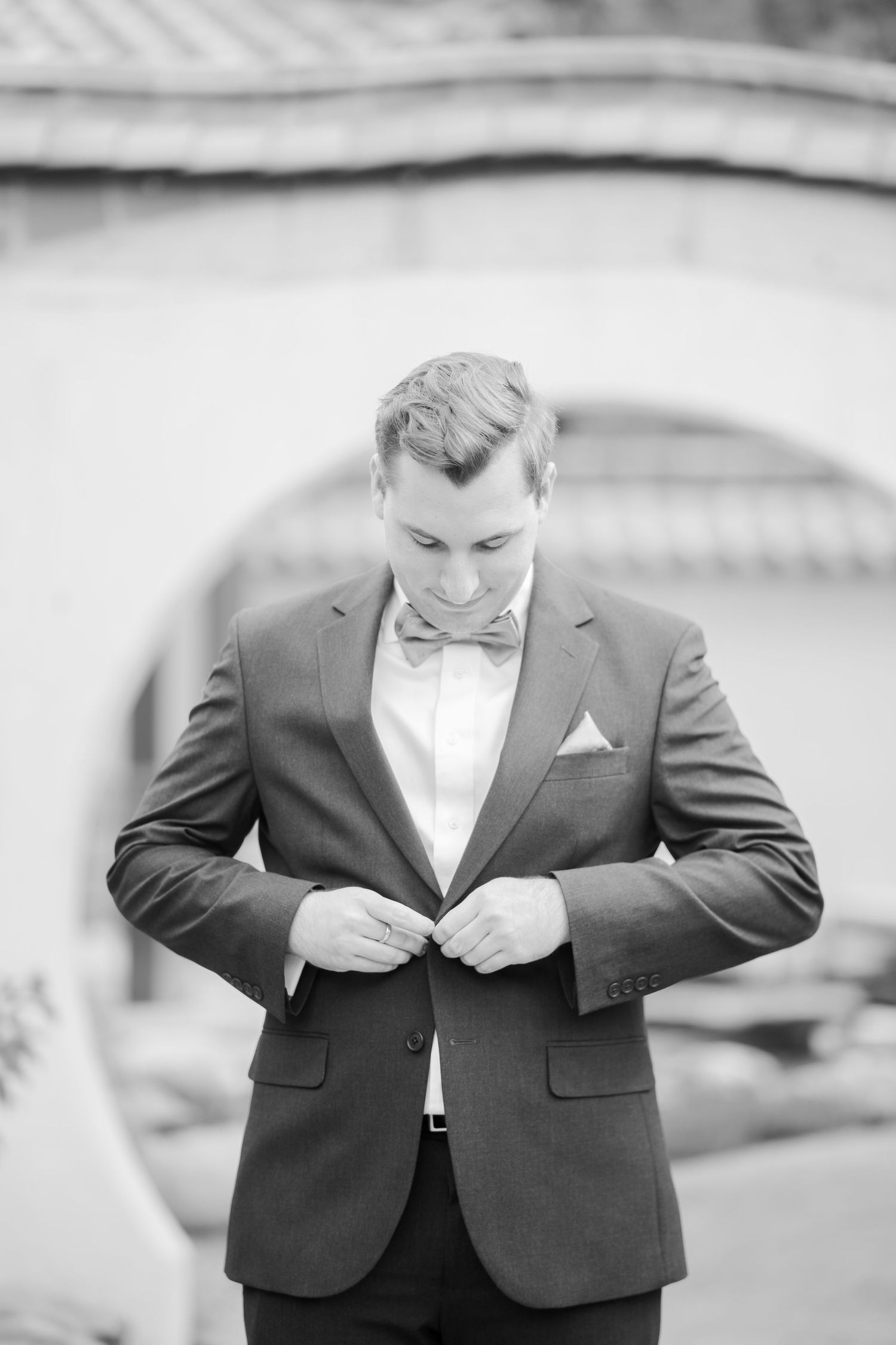 Groom in black suit and bow tie buttoning jacket in elegant black and white wedding portrait.