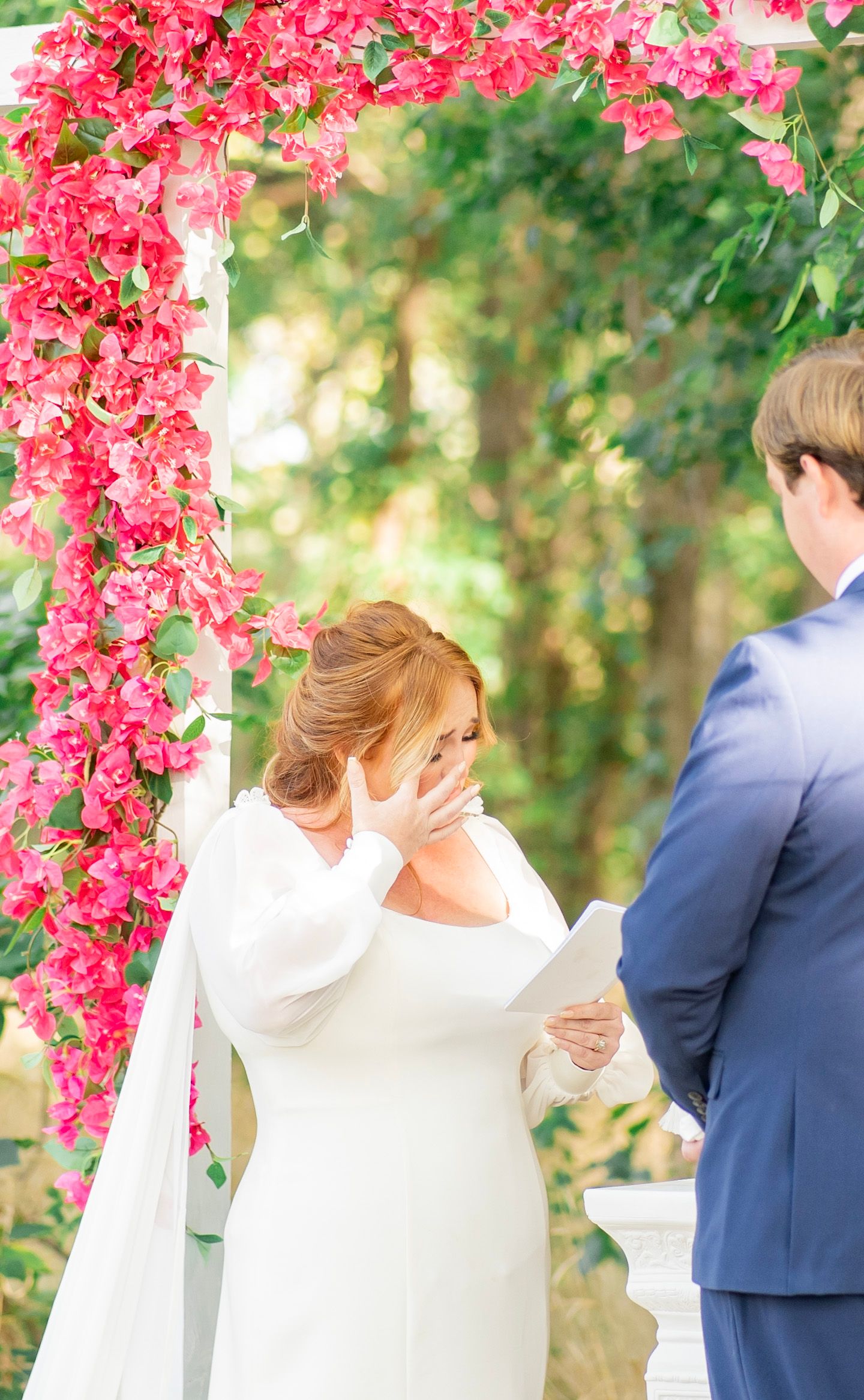 Emotional bride reading vows beneath stunning pink bougainvillea floral arch at outdoor garden wedding.