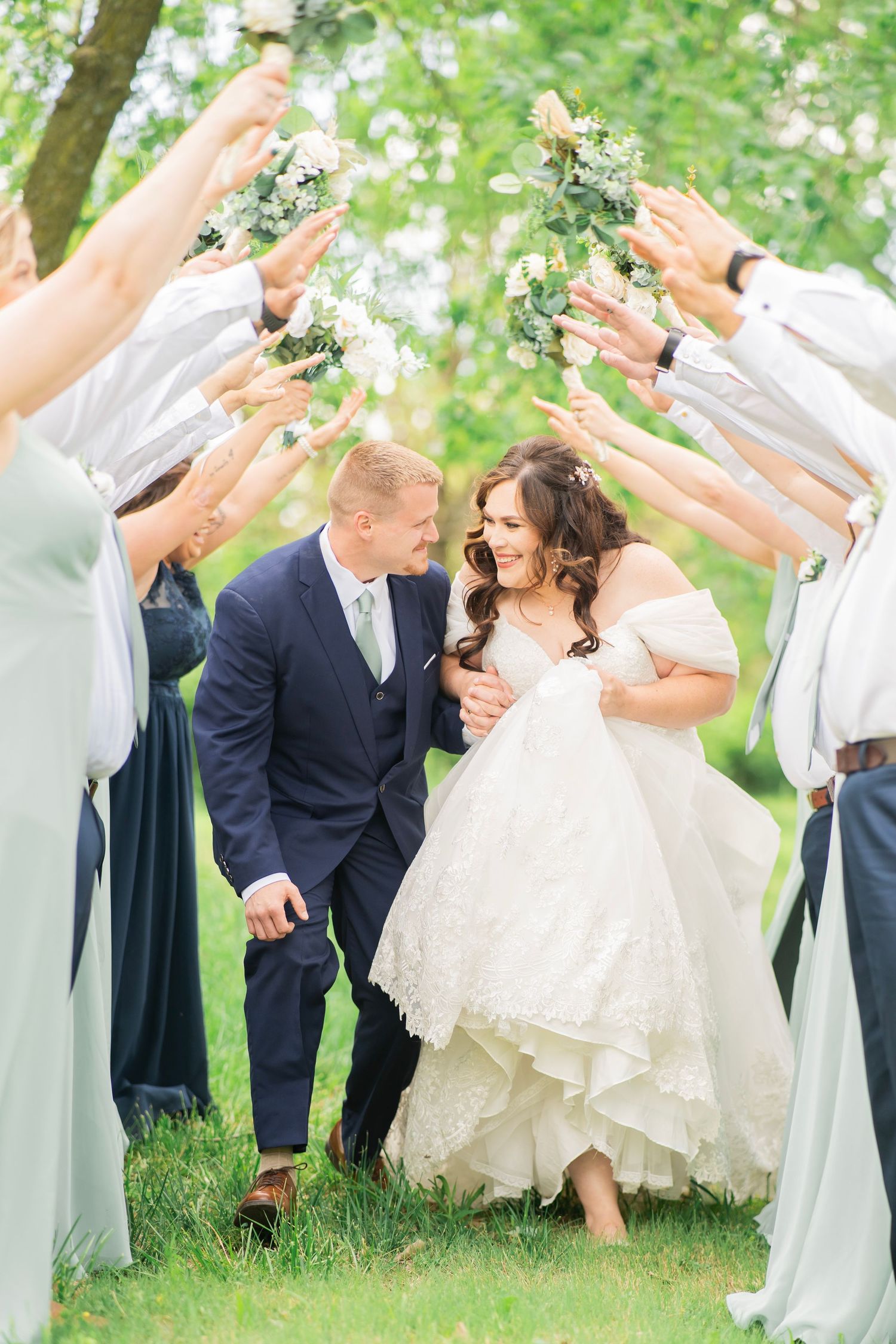 Bride and groom walk through a tunnel of raised bouquets held by their wedding party outdoors.
