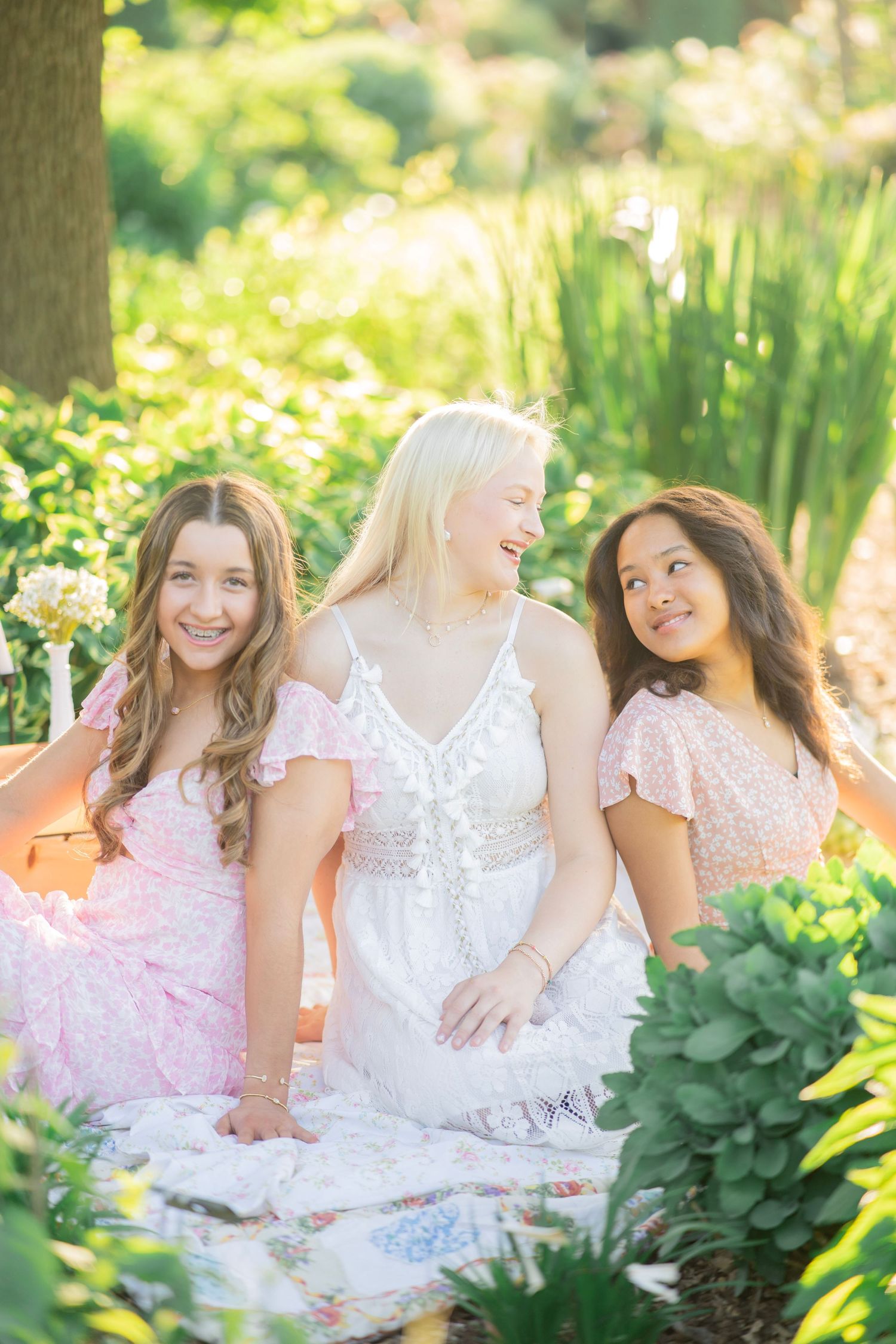 Three teenage girls in pink and white dresses sit together smiling in a sunlit garden setting.