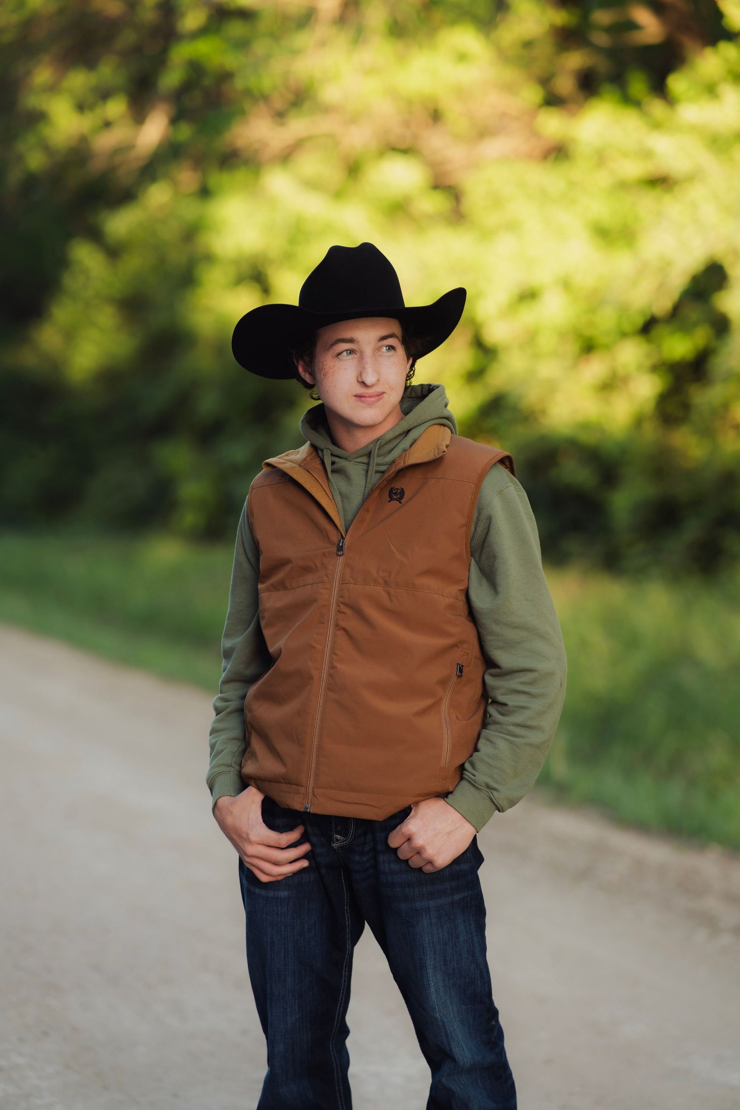 Young man in black cowboy hat and brown vest poses on a rural dirt road surrounded by lush green trees.