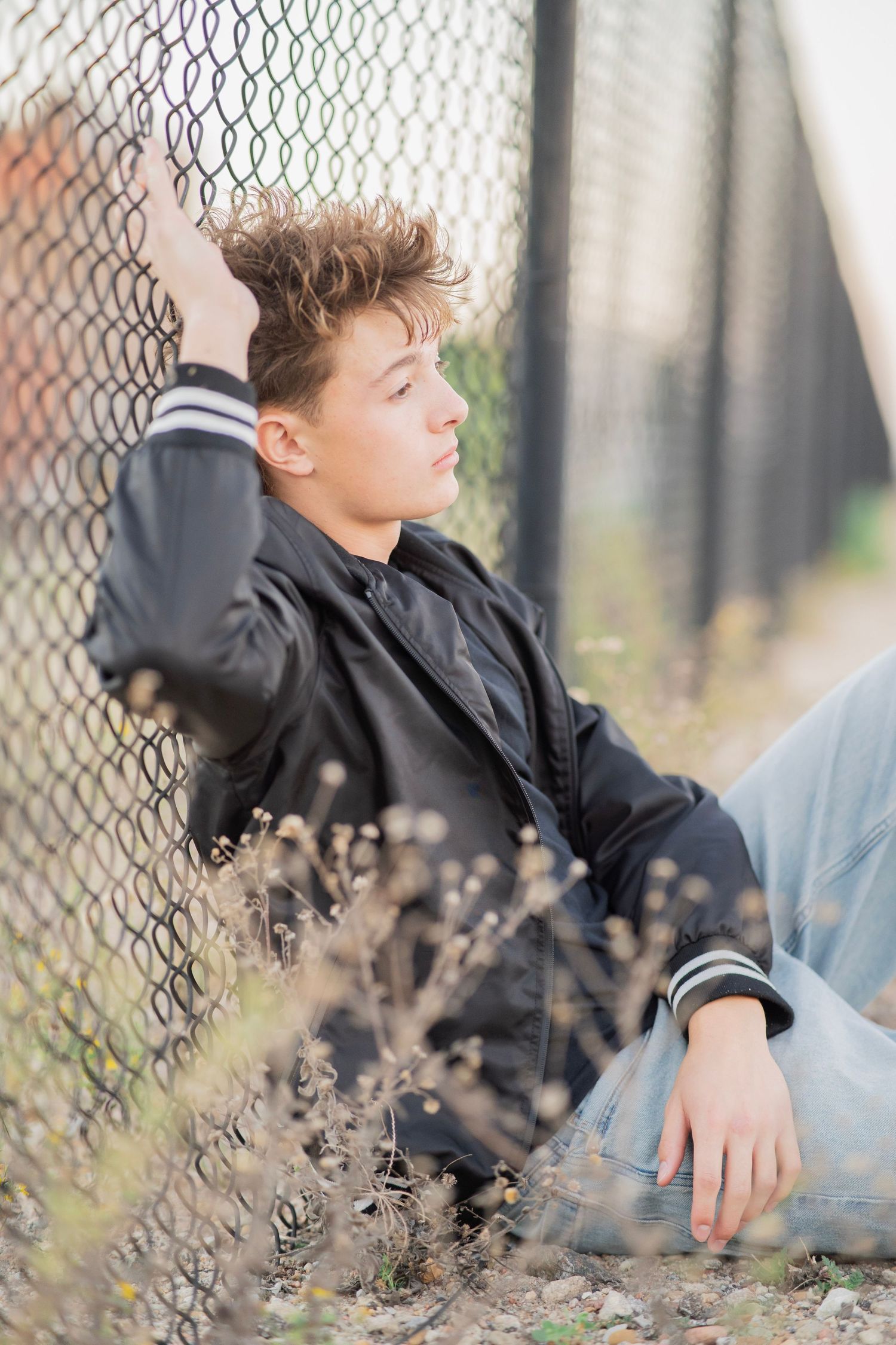 A teenage boy in a black bomber jacket and jeans sits against a chain-link fence outdoors, looking thoughtfully to the side.