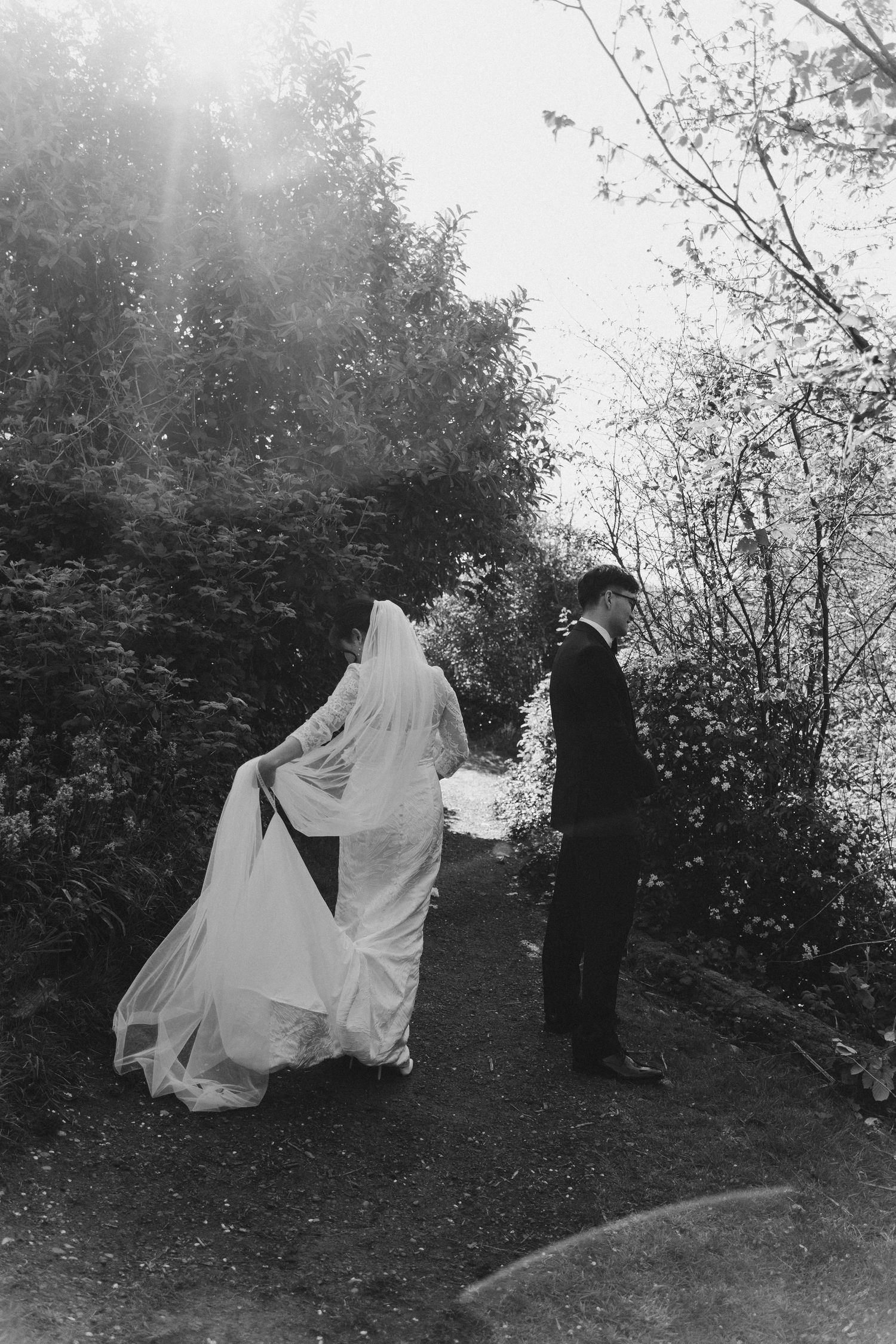 Black and white photo of bride and groom walking on a garden path, her veil flowing in the breeze.