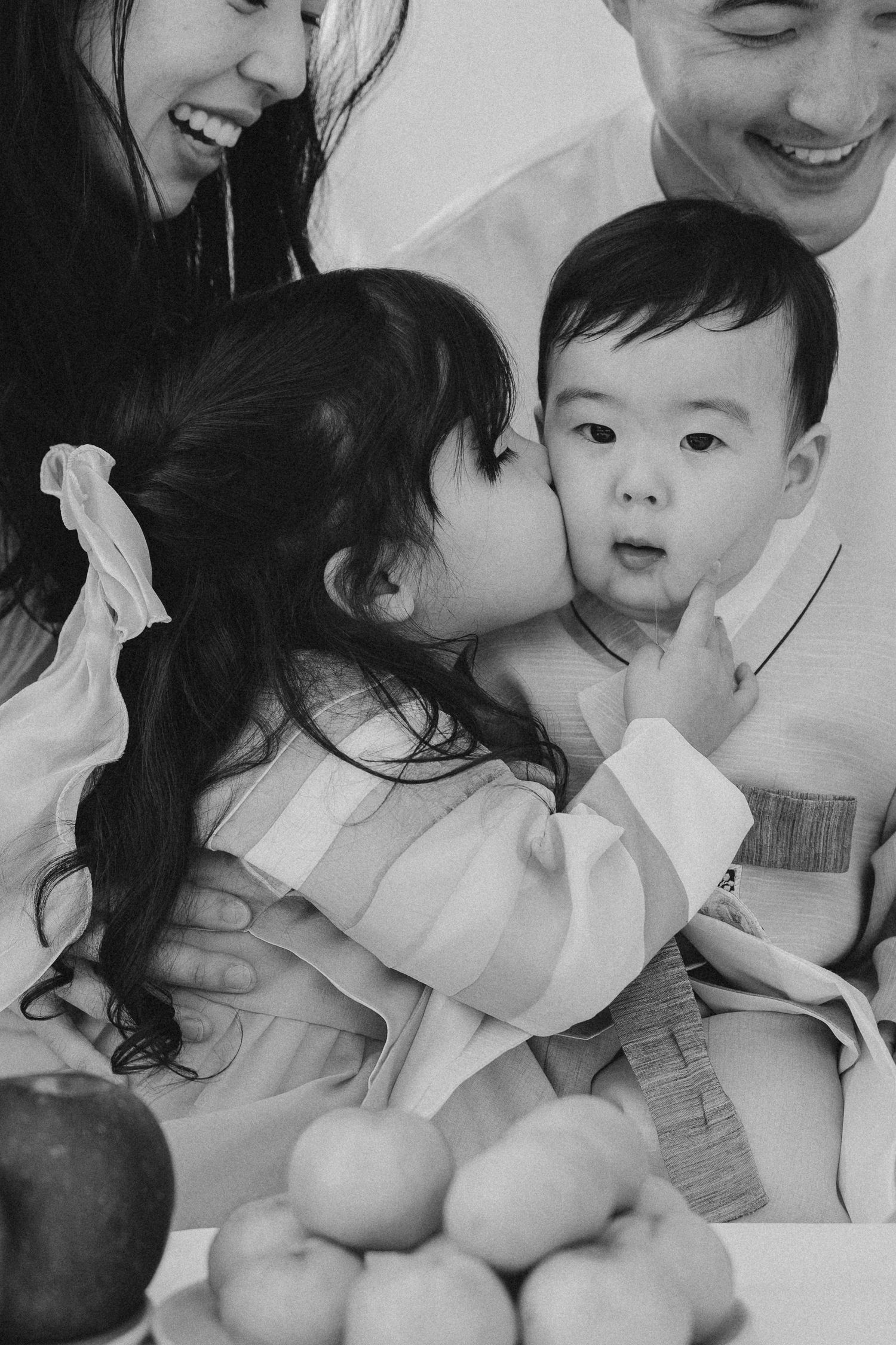Black and white photo of a young girl kissing a baby on the cheek, held by smiling parents in traditional Korean attire.