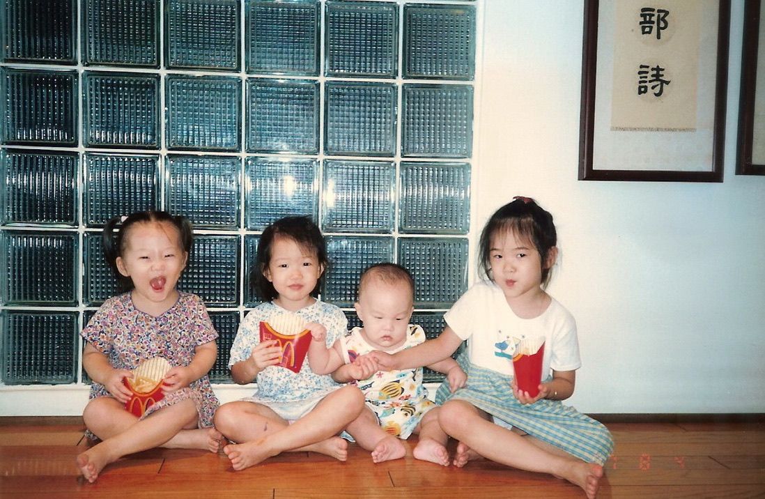 Three young children sit together on a wooden floor in what appears to be a dance or martial arts studio.
