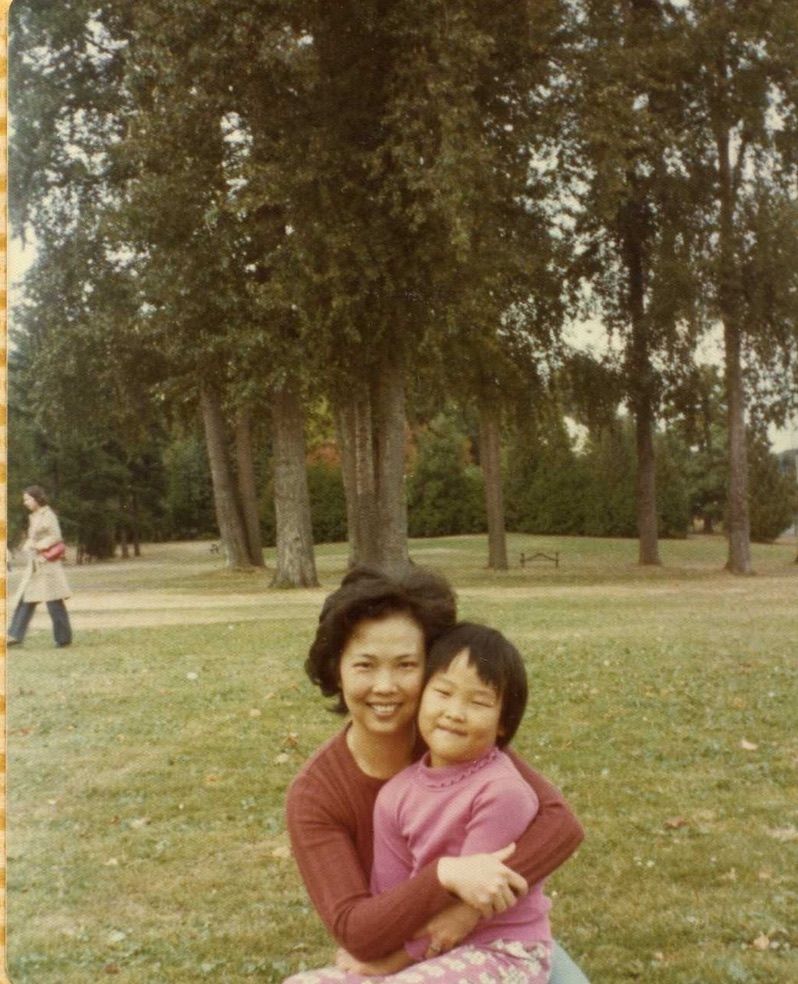 A parent and child share playful moments together on a grassy lawn surrounded by tall trees in a park setting.