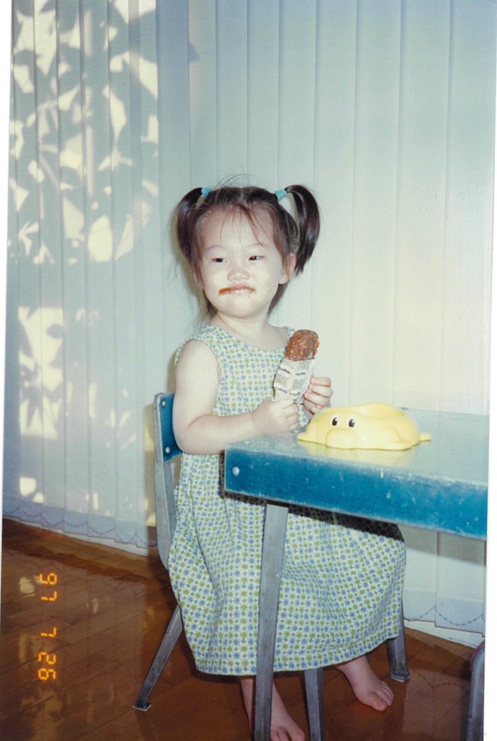 A young child in a patterned dress sits at a blue table with what appears to be a small cake or dessert.