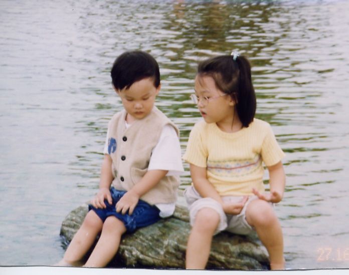 Two children sit by the water's edge with a small stuffed toy on a sunny day.