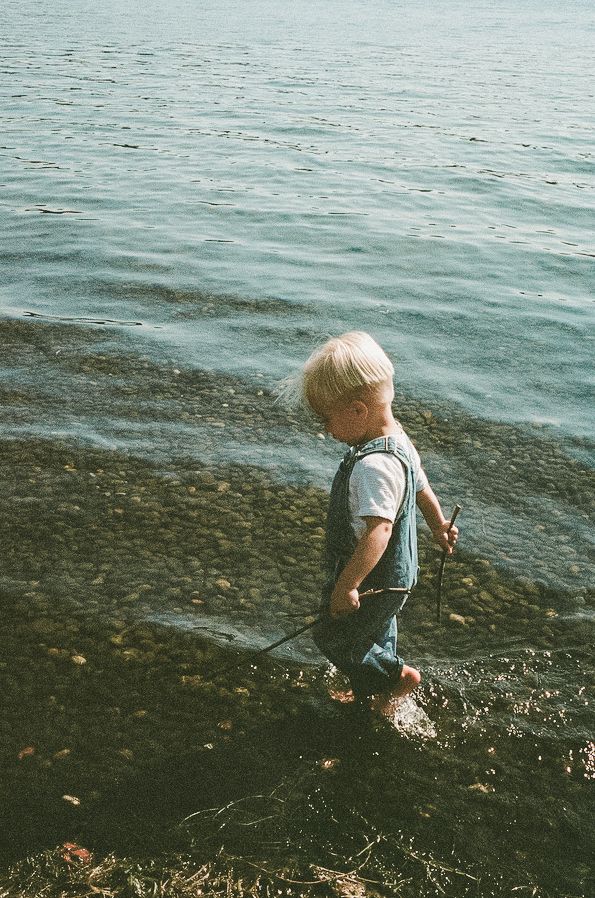 Blonde toddler in denim overalls playing at rocky shoreline, holding a stick near calm water.