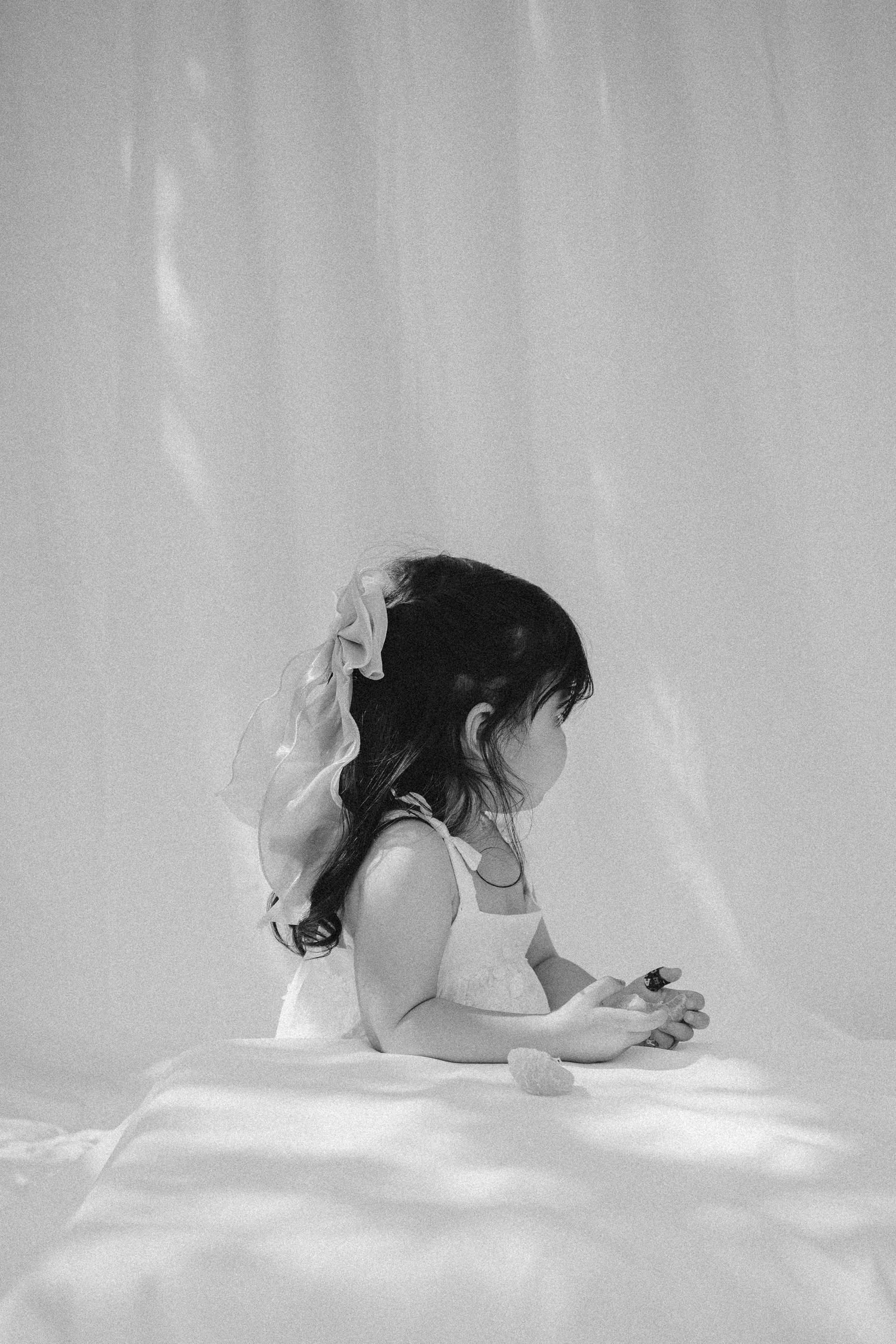 Black and white photo of a young girl in a white dress with a bow in her hair, sitting quietly in soft light.