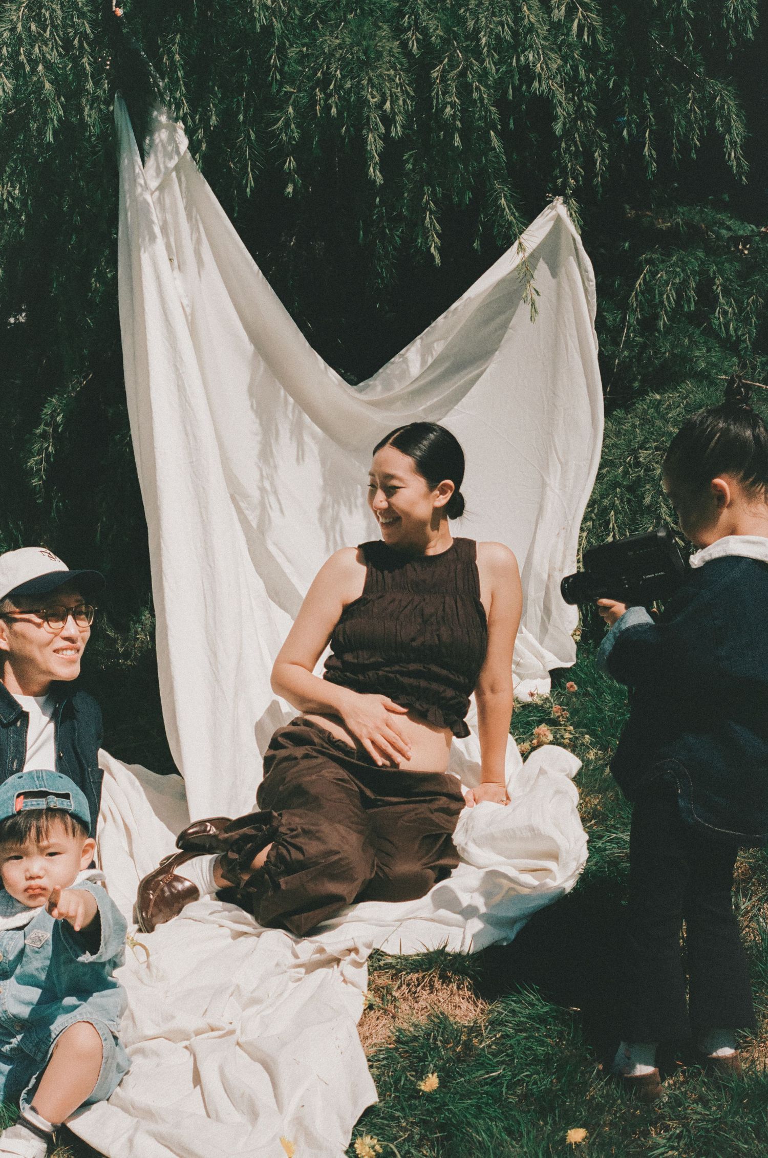 Pregnant woman in dark dress sits on white blanket outdoors with family, willow tree and white fabric backdrop behind her.