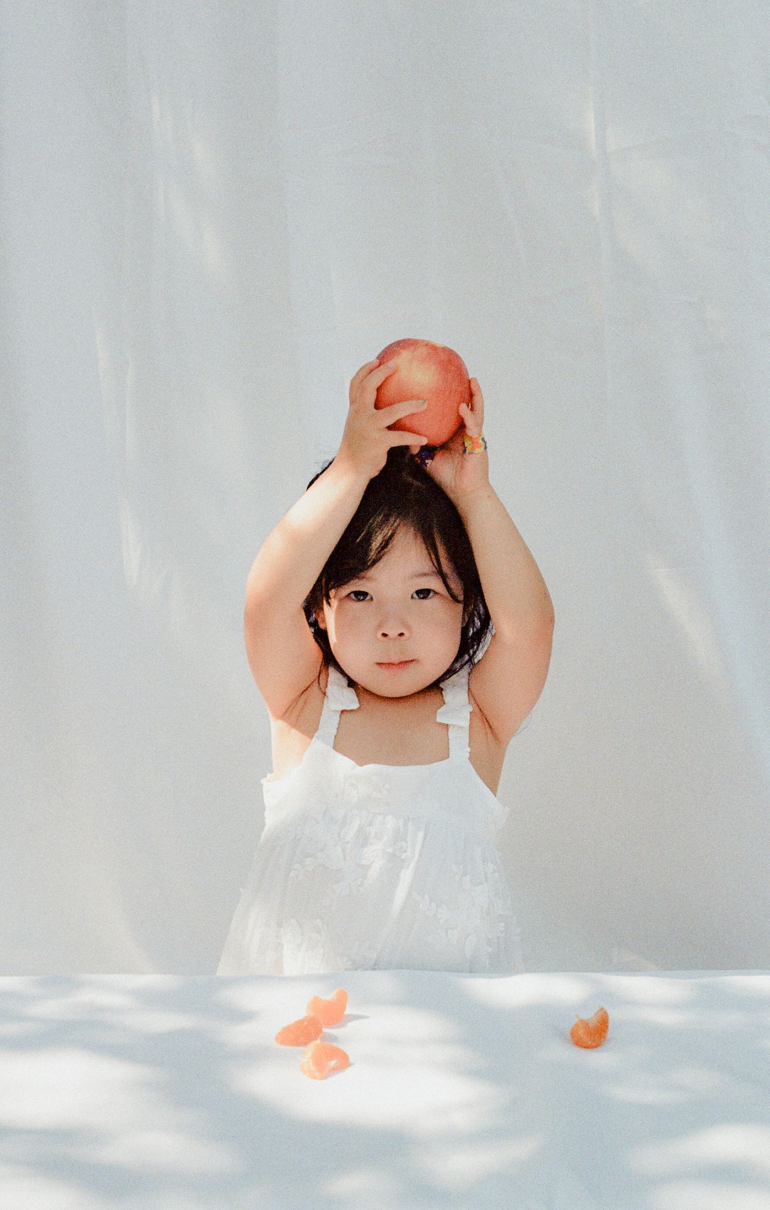 A young girl in a white dress holds a red apple above her head at a white table with orange peels.