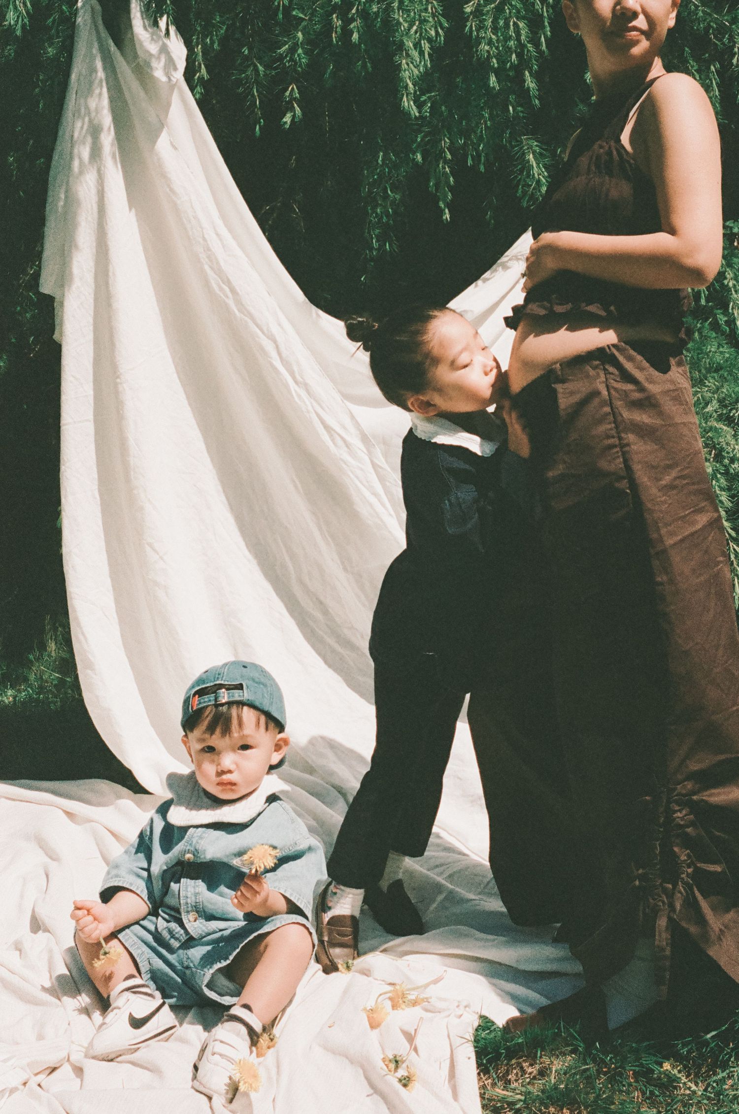 A young boy sits on a white blanket outdoors while another child kisses a woman in a brown dress near a willow tree.