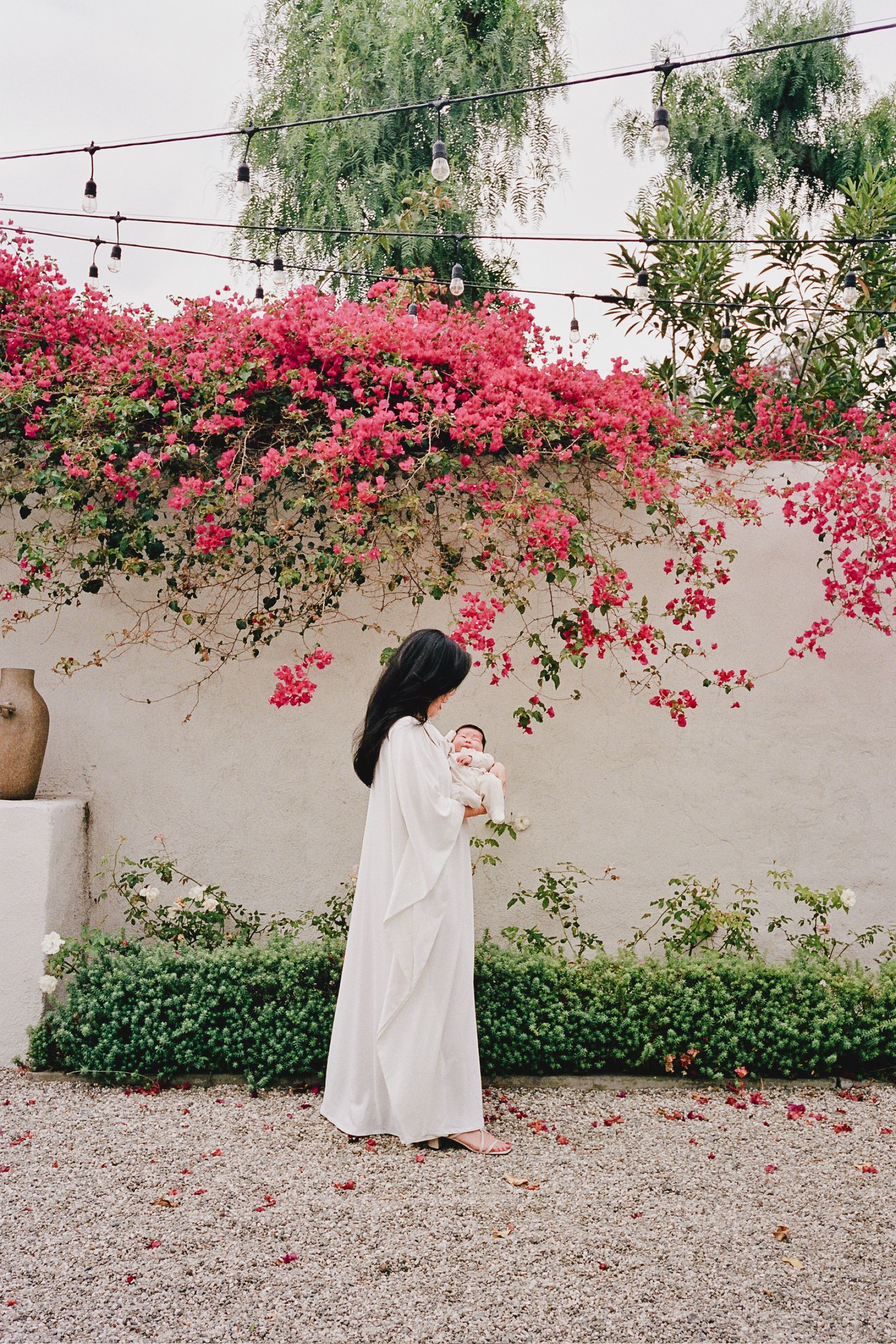 A woman in a white dress holds a newborn baby in front of a wall covered in vibrant pink bougainvillea flowers.