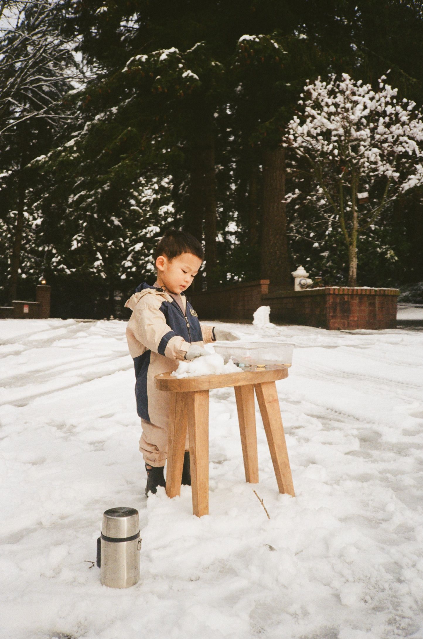A child sits at a small wooden bench in a snowy winter landscape with a thermos nearby.