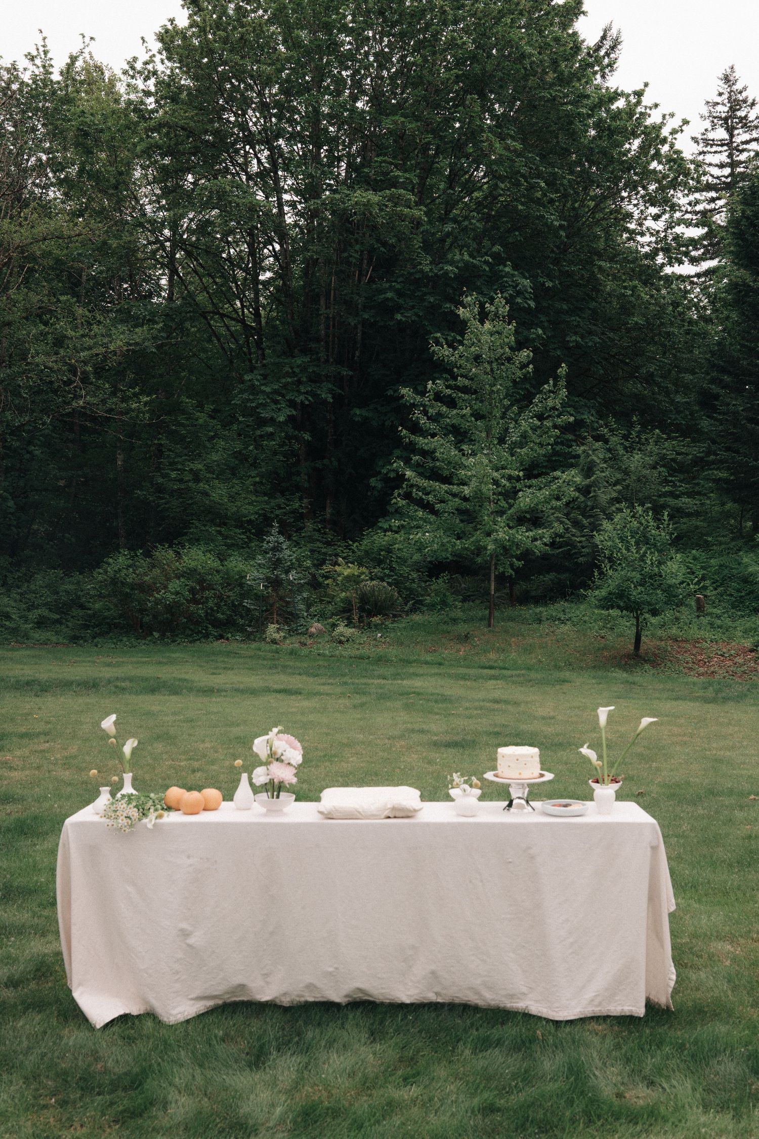 Elegant outdoor wedding cake table with white linen, floral arrangements, and a tiered cake set against a lush green forest backdrop.