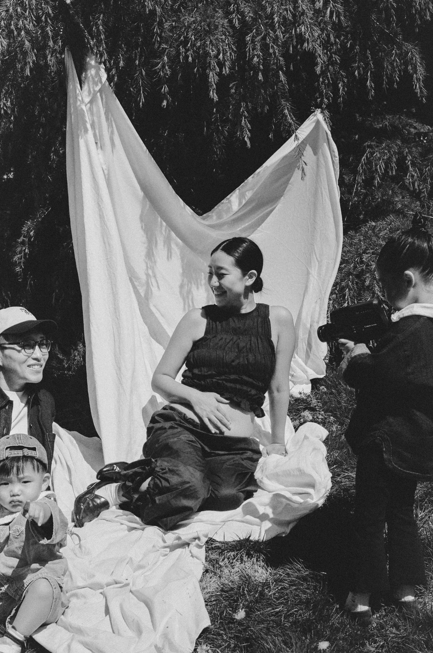 Black and white photo of a woman sitting outdoors under a white fabric backdrop during a maternity photoshoot.