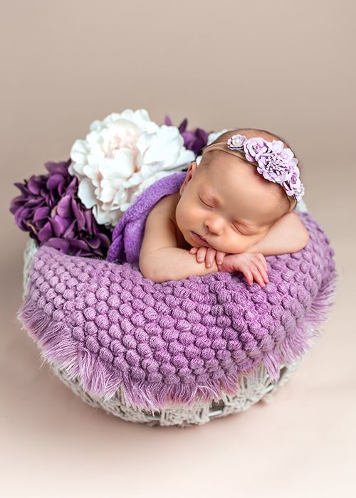 Newborn photo of a baby nestled in a purple knitted blanket with flowers in a decorative white basket.