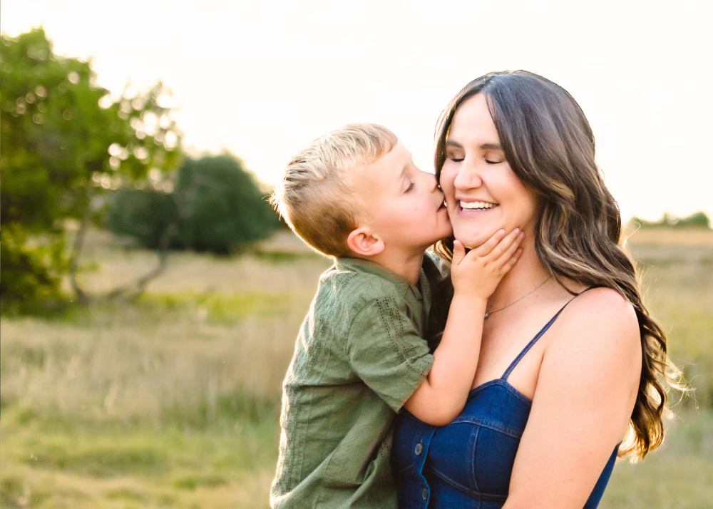 A tender moment captured between mother and child with a kiss on the cheek in a sunny outdoor field setting in Colorado during family photos