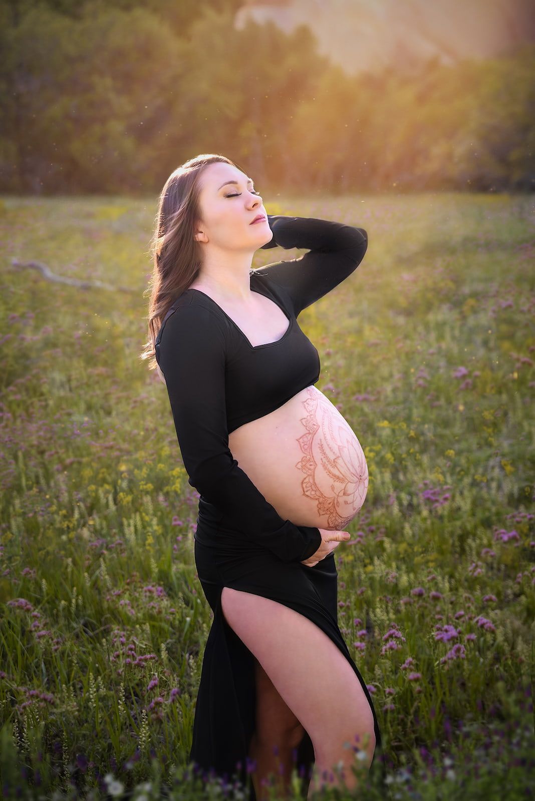 Artistic maternity photo of an expectant mother in black dress with henna belly art standing in wildflower meadow at sunset.
