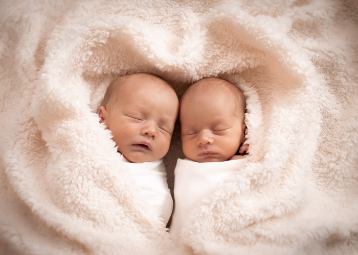 Newborn photography of twins swaddled together in soft beige blankets forming a heart shape around them.