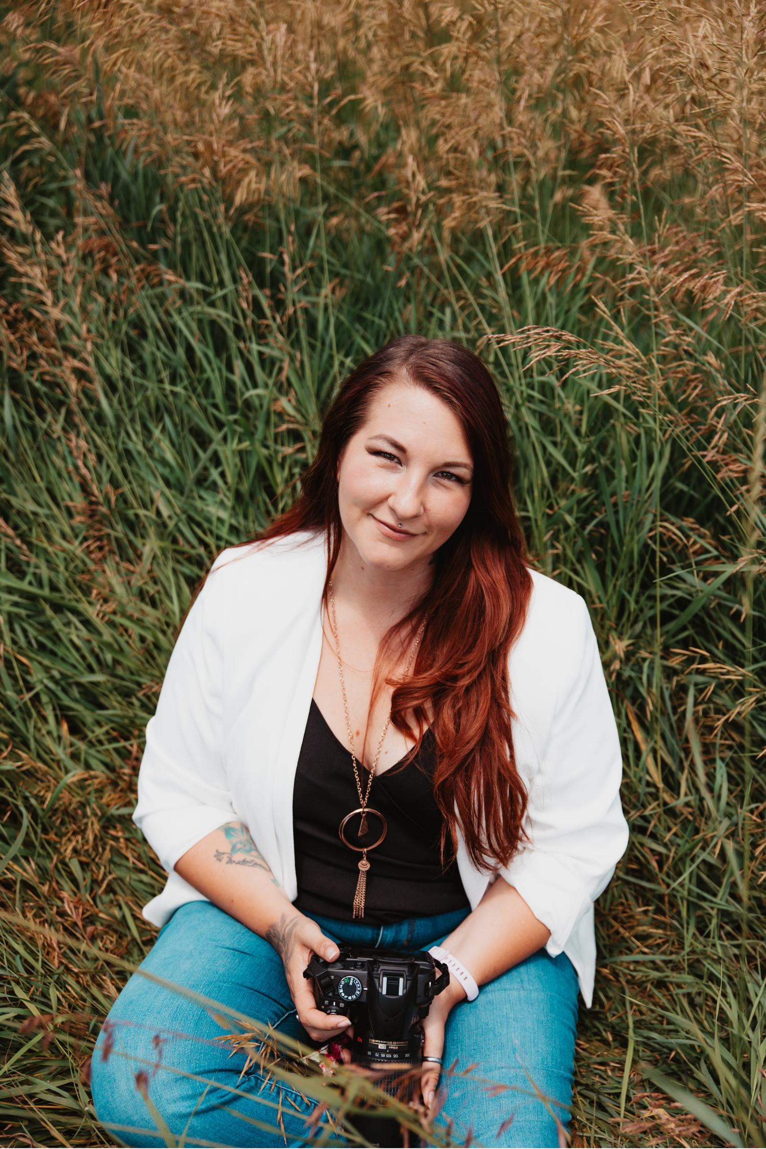 Portrait of photographer sitting in tall grass holding camera with white blazer and jeans.
