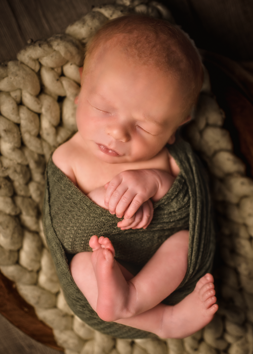 Newborn baby posed peacefully wrapped in olive green fabric on chunky knit blanket in warm studio lighting for newborn photos.
