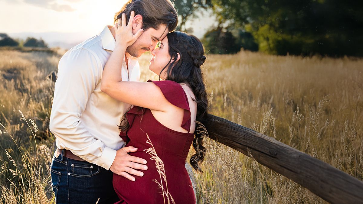 A couple embraces in a golden field during sunset while wearing burgundy and white outfits for their engagement photos.