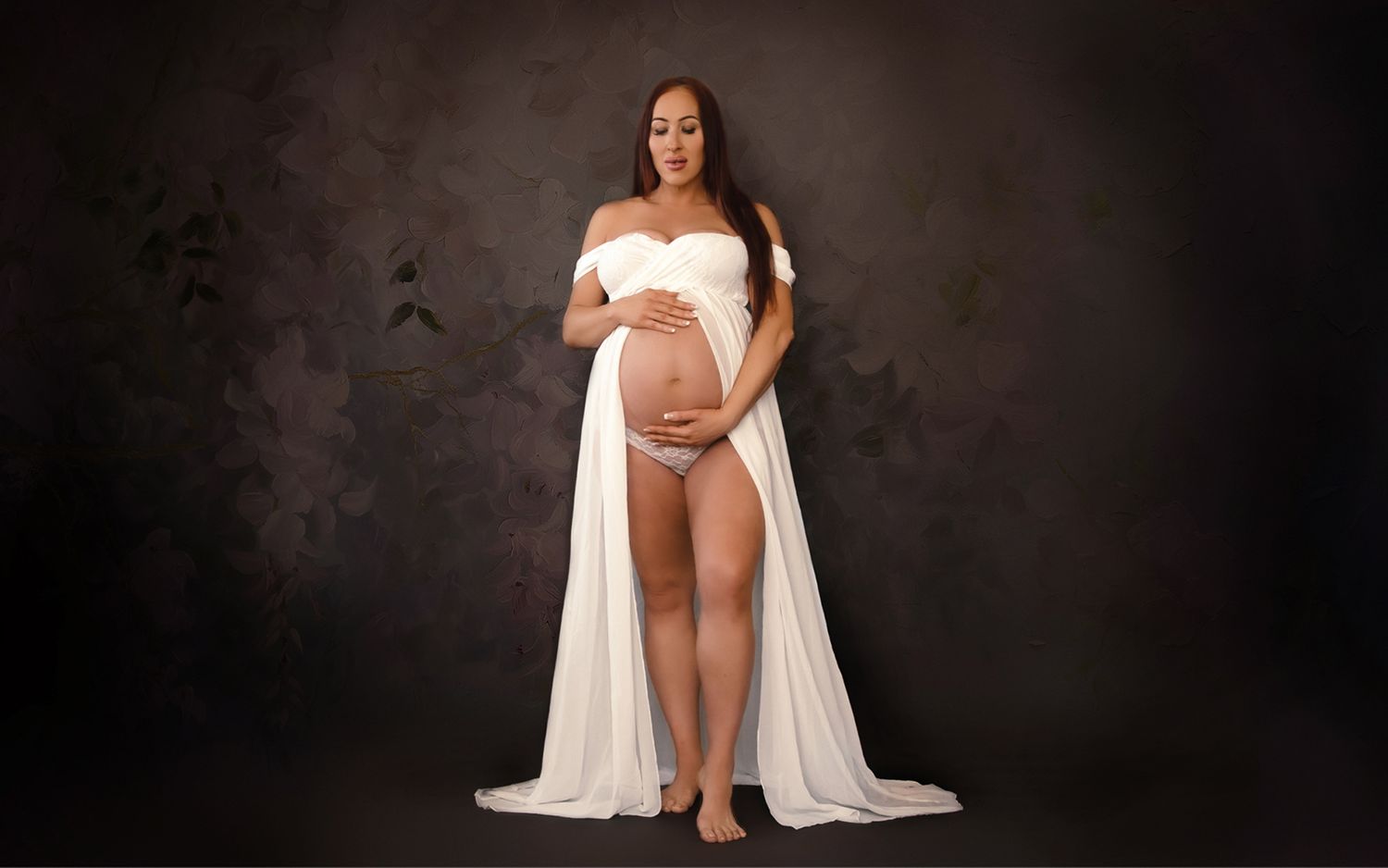 Elegant studio maternity portrait in flowing white gown against floral backdrop by Denver maternity photographer.