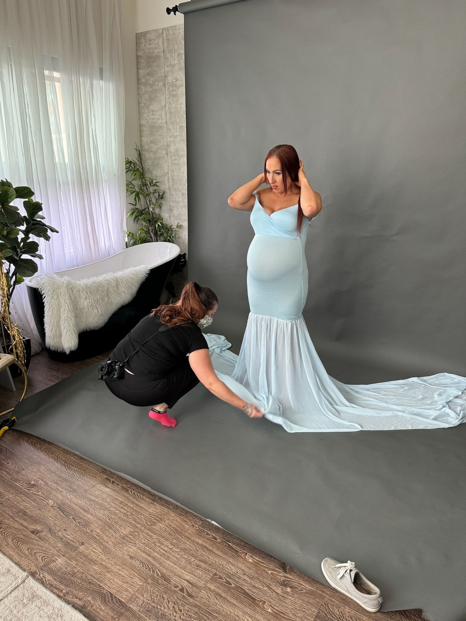 Behind the scenes photo of maternity photoshoot with blue dress being adjusted on grey backdrop.
